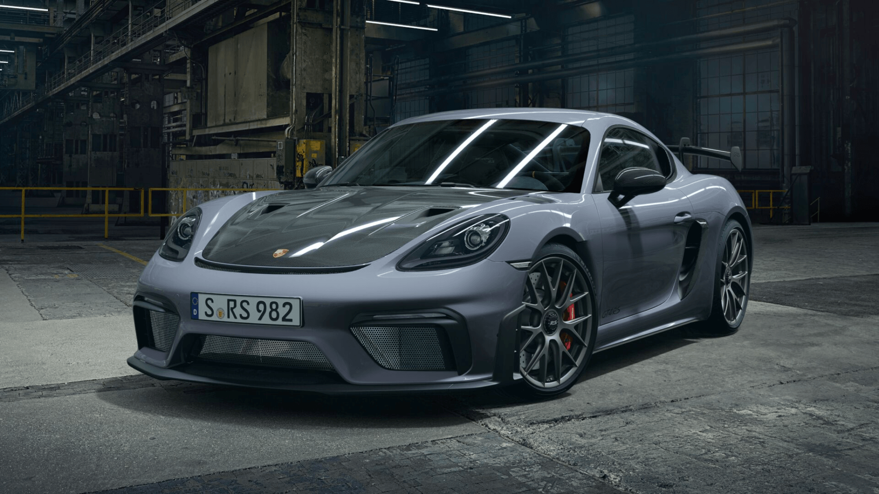 A lavender-grey Porsche Cayman GT4 RS sports car, featuring a dark carbon fiber hood, parked indoors in a dimly lit, industrial-style garage or factory.