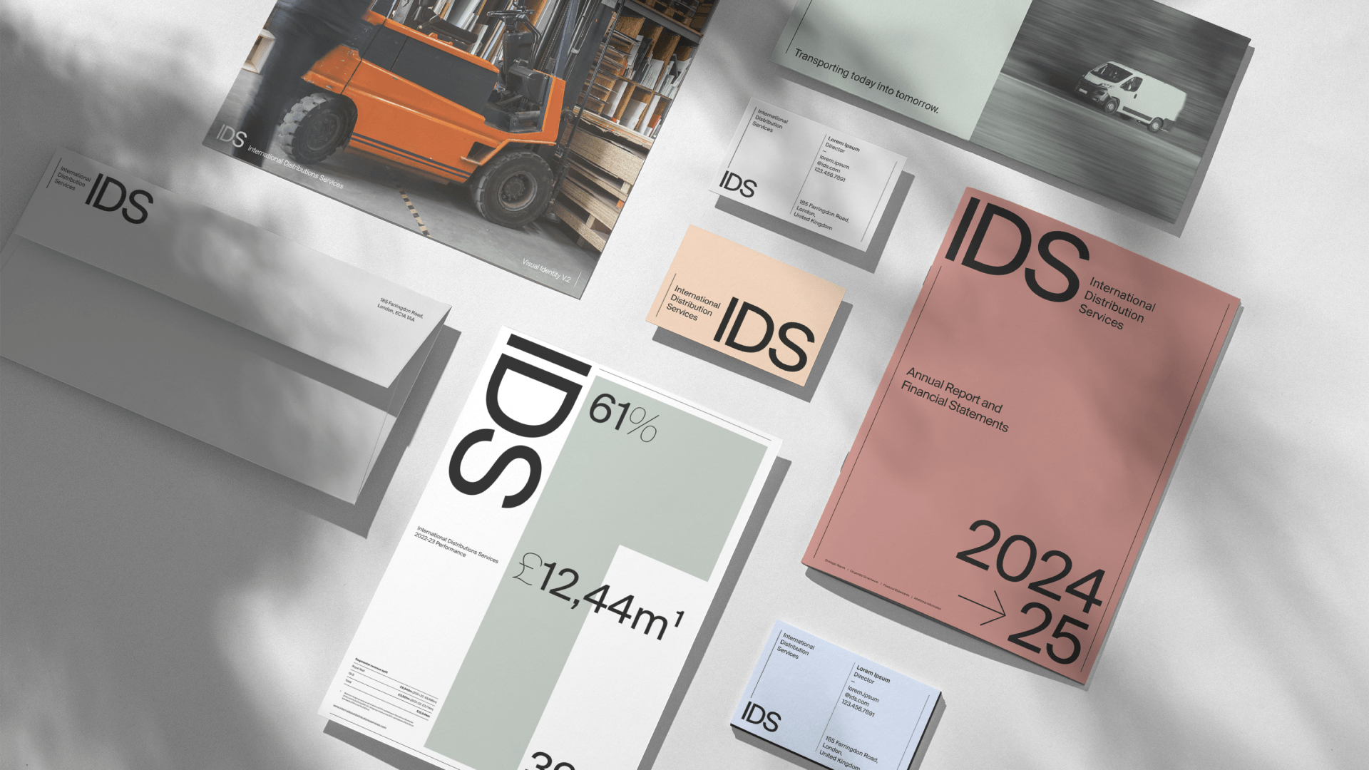 Flat lay of corporate branding materials for "IDS International Distribution Services," including reports, business cards, and an envelope, featuring a modern design with black, white, grey, and salmon pink typography and a partial image of an orange forklift.