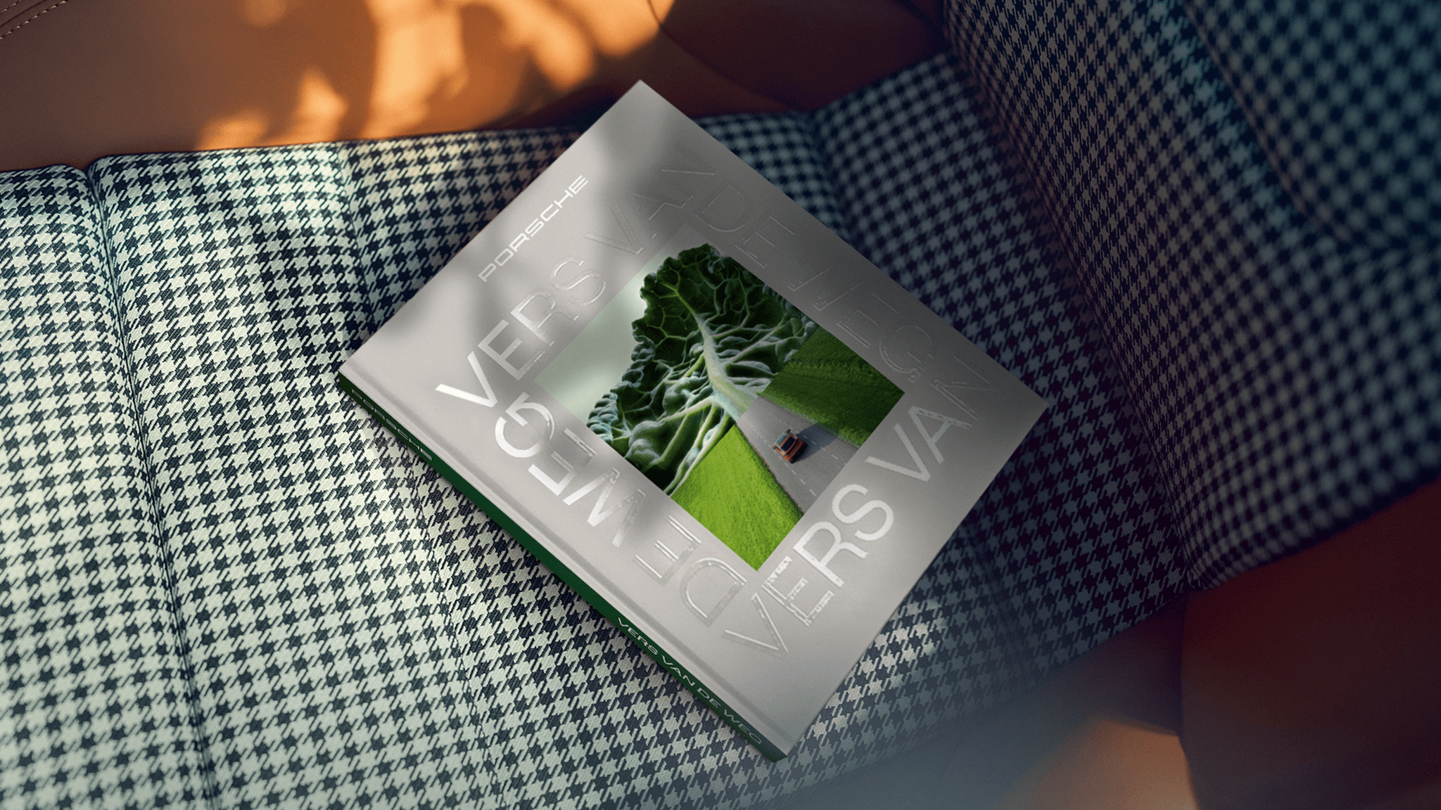 A square book or magazine titled "PORSCHE VERS VAN DE WEG" in large, glossy white lettering, resting on a houndstooth fabric background. The cover features a cutout or picture window showing a large piece of green kale or lettuce leaf blending into a patch of green grass with a small red Porsche in the distance.