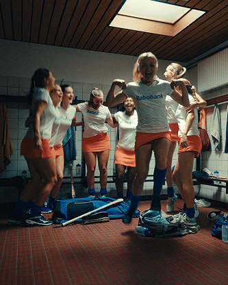 Cheering women's field hockey team in locker room, wearing white "Rabobank" shirts and orange skirts, gear and sticks on the floor.