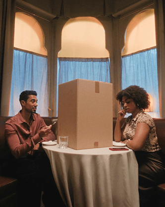 A man and a woman sit at a small restaurant table, separated by a large, tall cardboard box placed right in the center