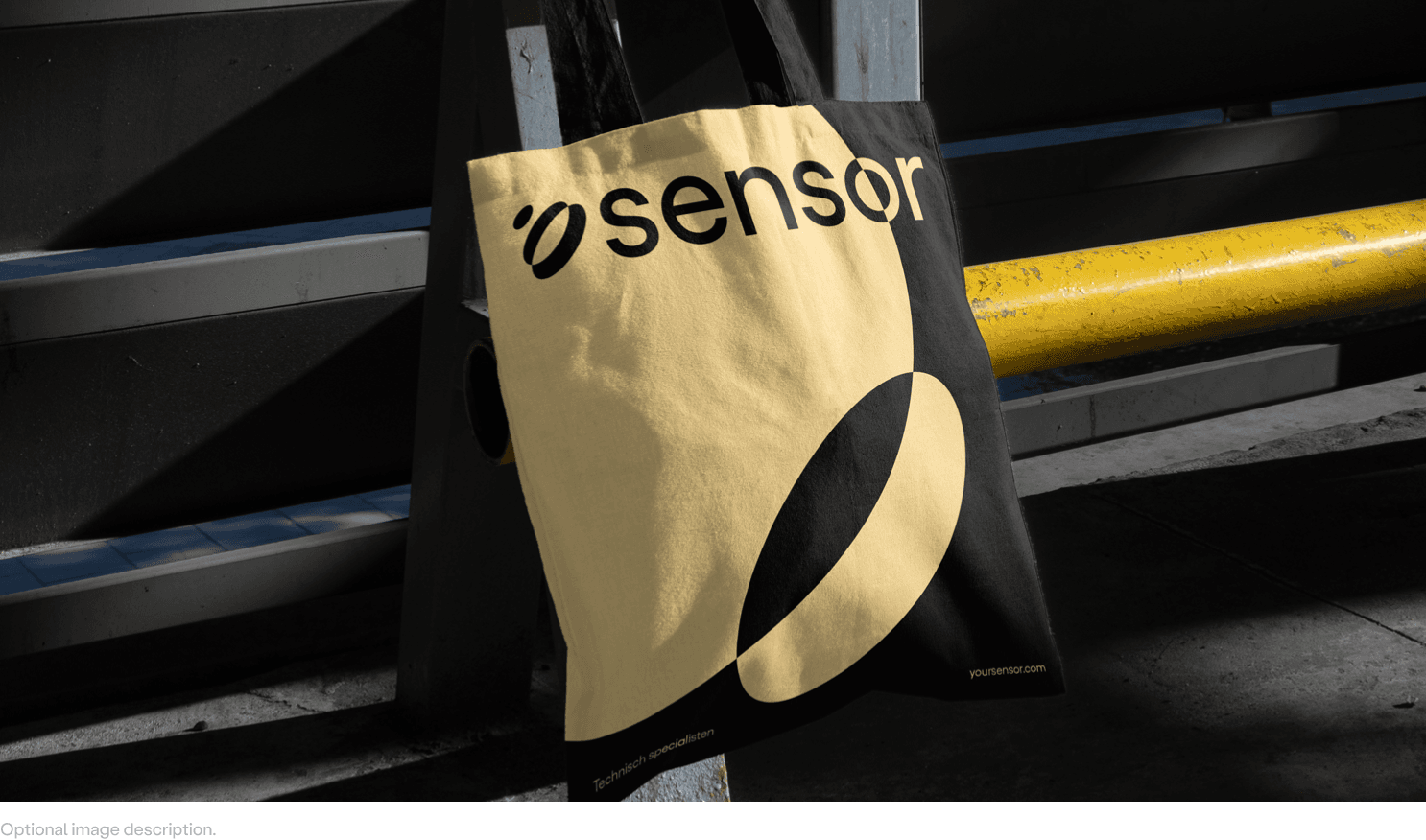 Yellow and black tote bag printed with 'sensor' logo hanging on a gray metal railing beside a yellow pipe.