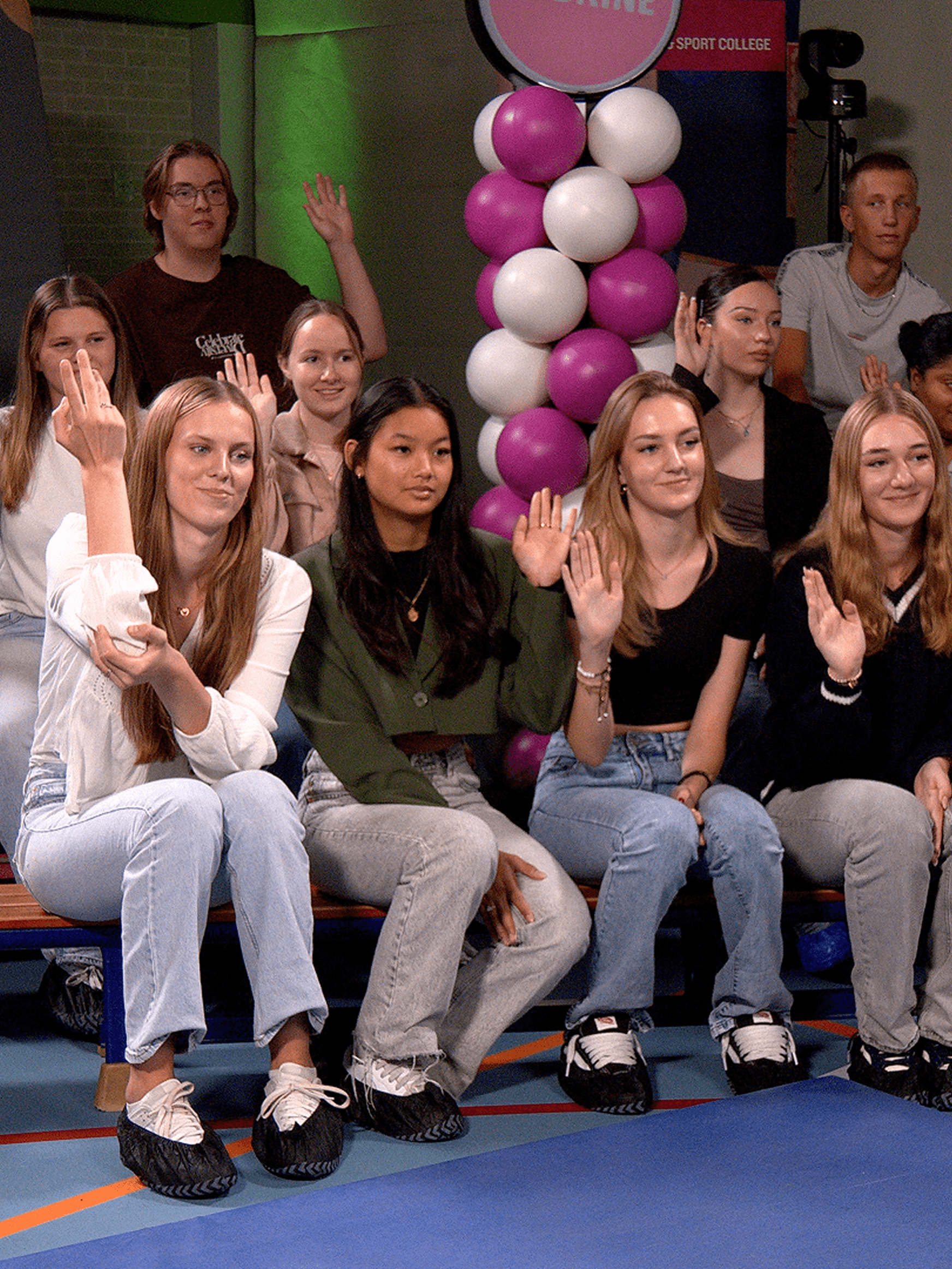 Smiling teens seated indoors, several raising hands next to a purple-and-white balloon column.