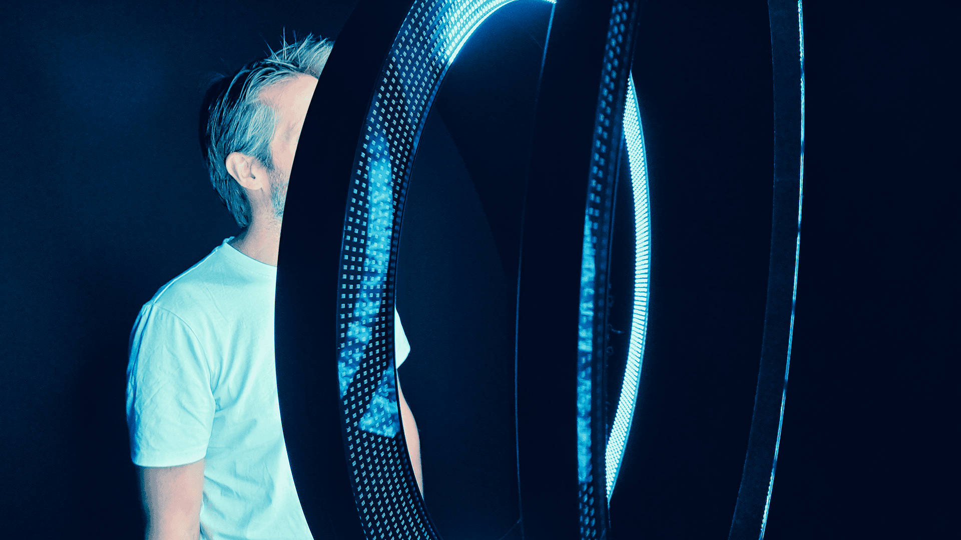 Person in a white T-shirt partially obscured by large glowing blue LED rings against a dark background.