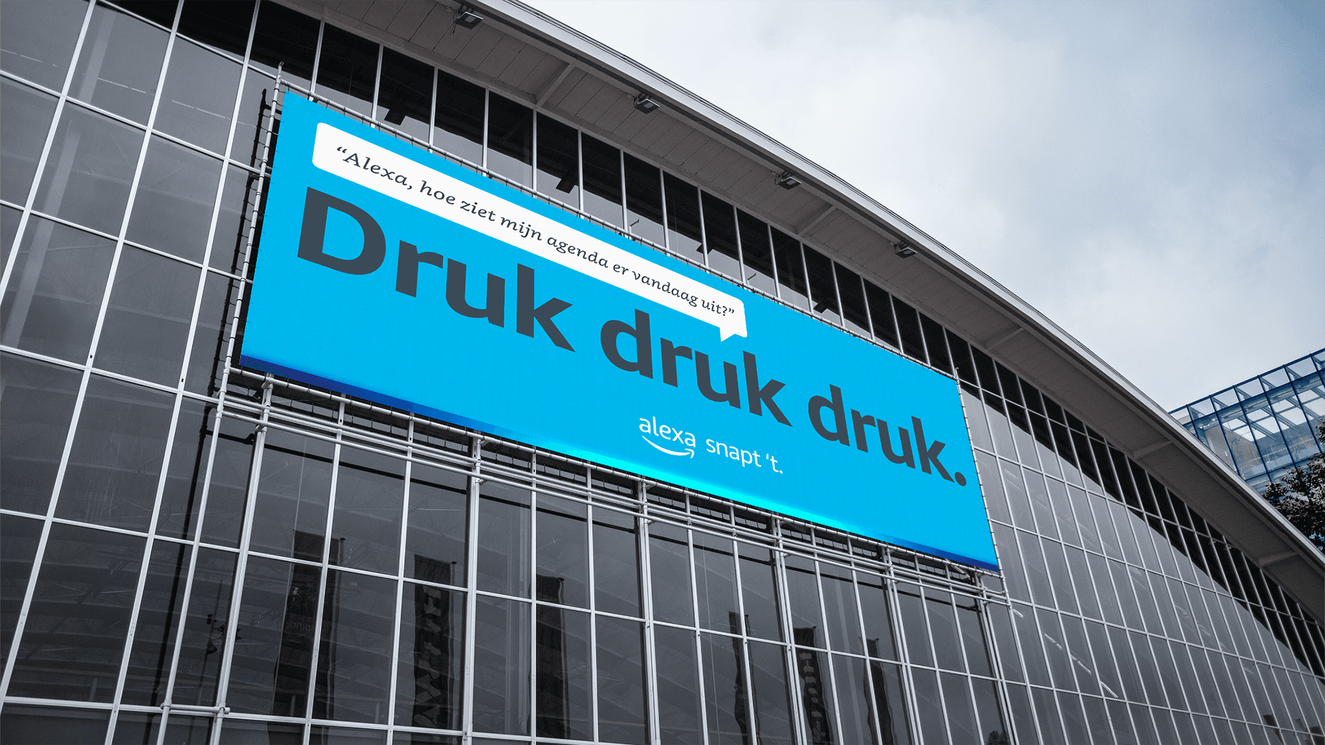 Bright blue billboard reading "Druk druk druk." mounted on a curved glass office façade, with small Alexa branding below.
