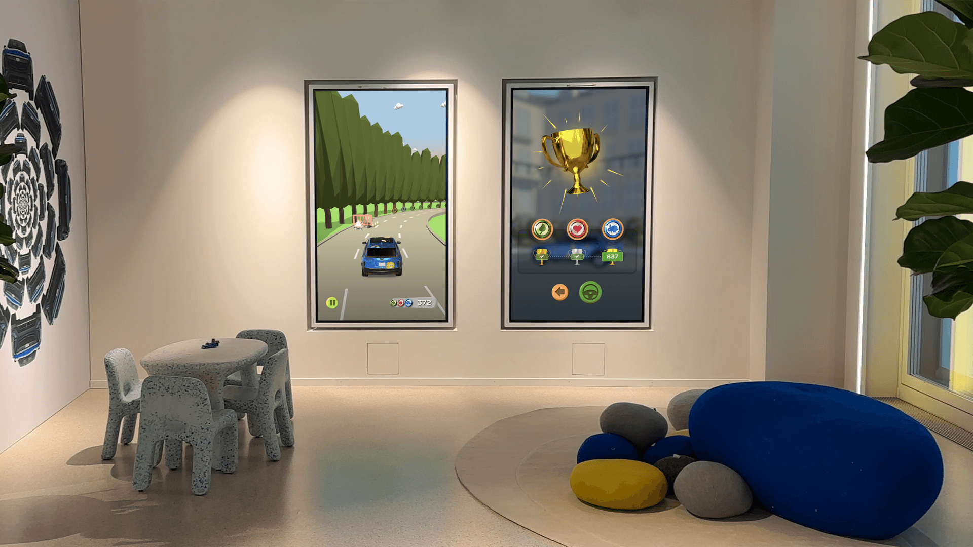 Playroom with two vertical game screens (racing and trophy UI), child-sized table and chairs, blue pebble seating and a potted plant.