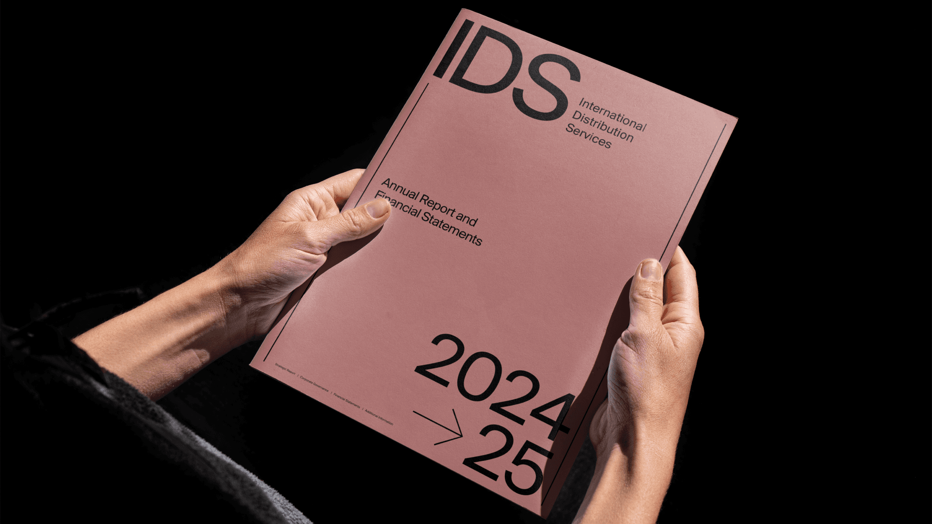 Two hands hold a pink IDS annual report and financial statements booklet dated 2024–25 against a black background.