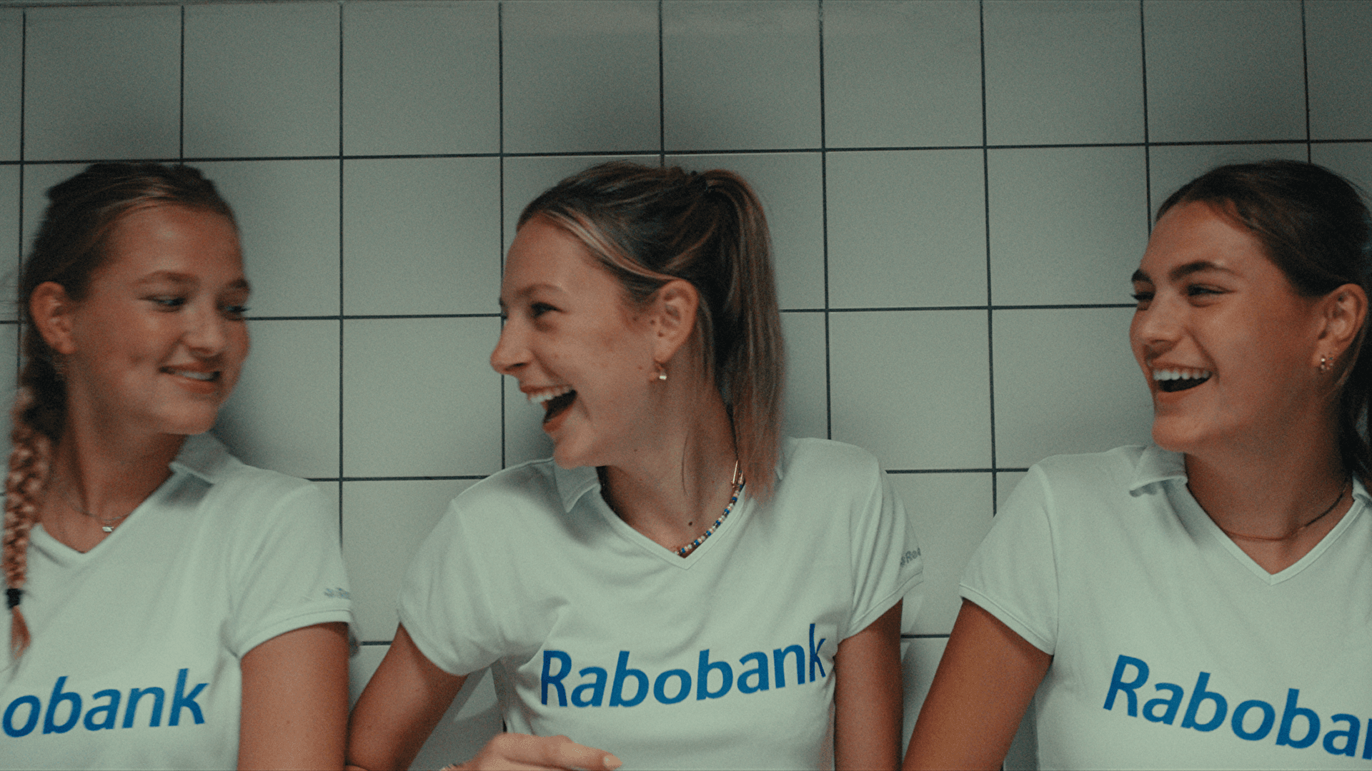 Three young women in white Rabobank shirts laughing together in front of a tiled wall.