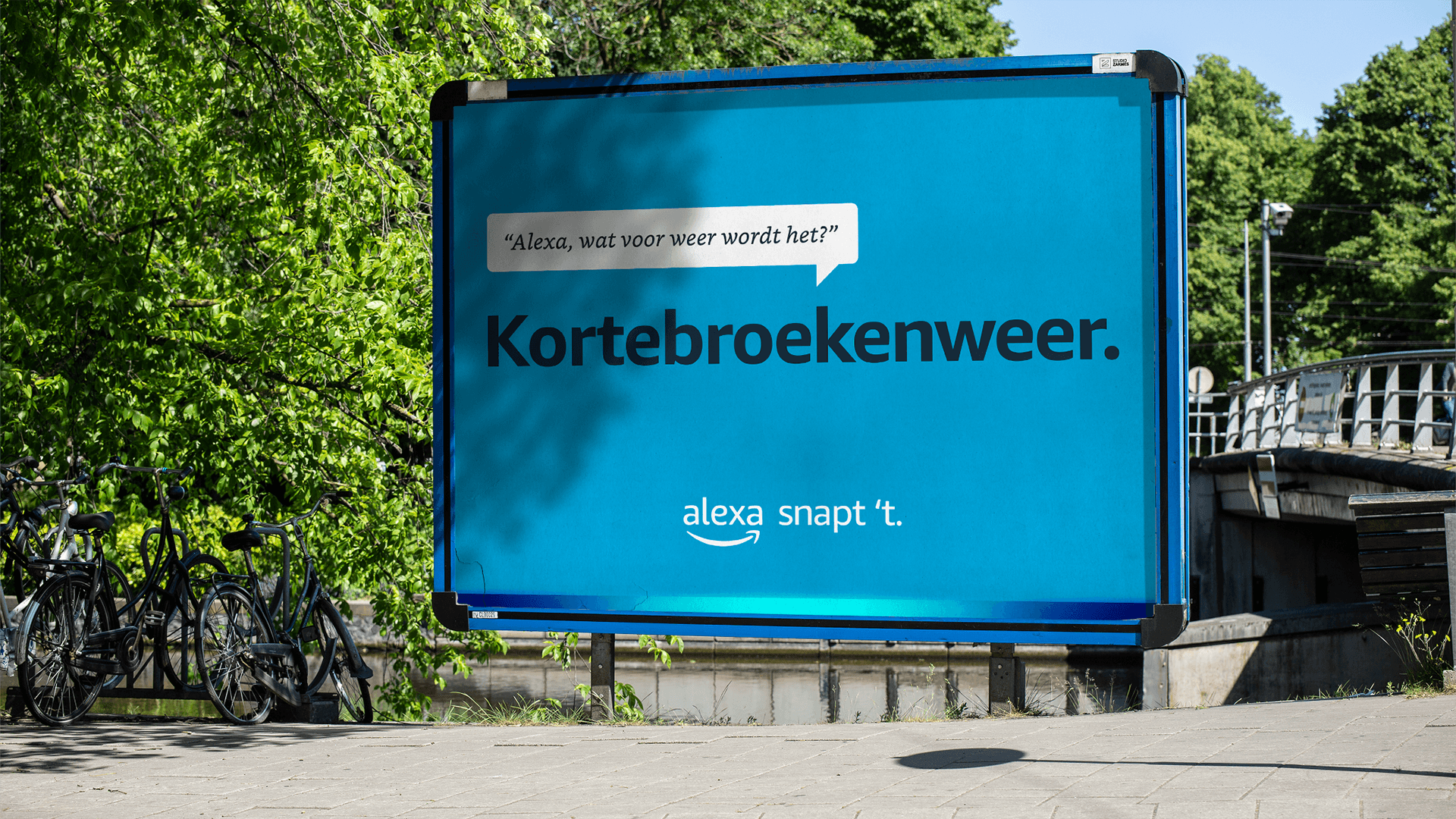 Blue outdoor billboard reading "Kortebroekenweer." with Alexa logo; bicycles parked nearby, trees and a bridge in the background.