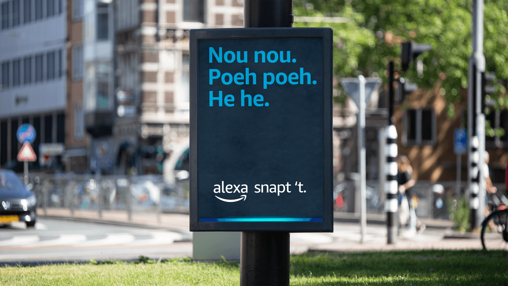 Lamppost ad panel reading "Nou nou. Poeh poeh. He he." and "alexa snapt 't." with Amazon smile logo, city street scene.