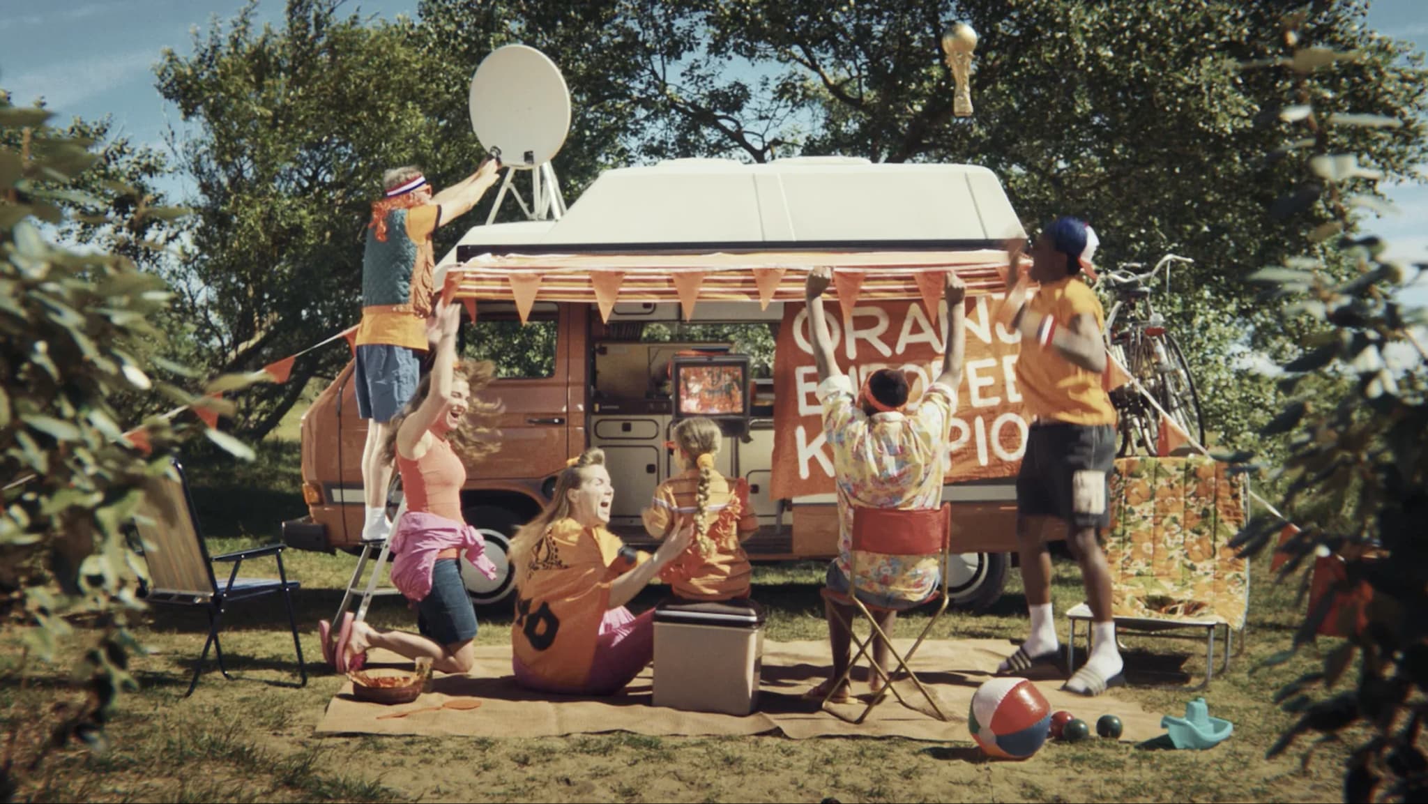 A group of fans dressed in orange "Oranje" gear celebrate outside an orange Volkswagen camper van equipped with a satellite dish, watching a small television during a picnic.