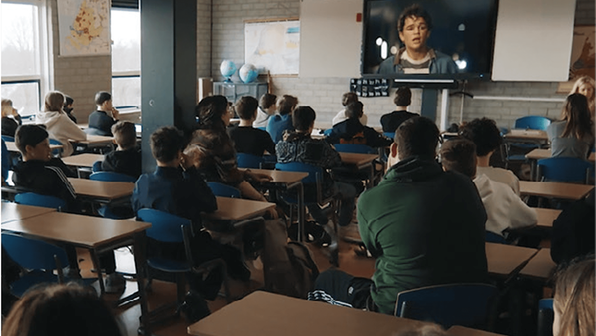 High school classroom with students seated at desks watching a film on a large screen at the front of the room.