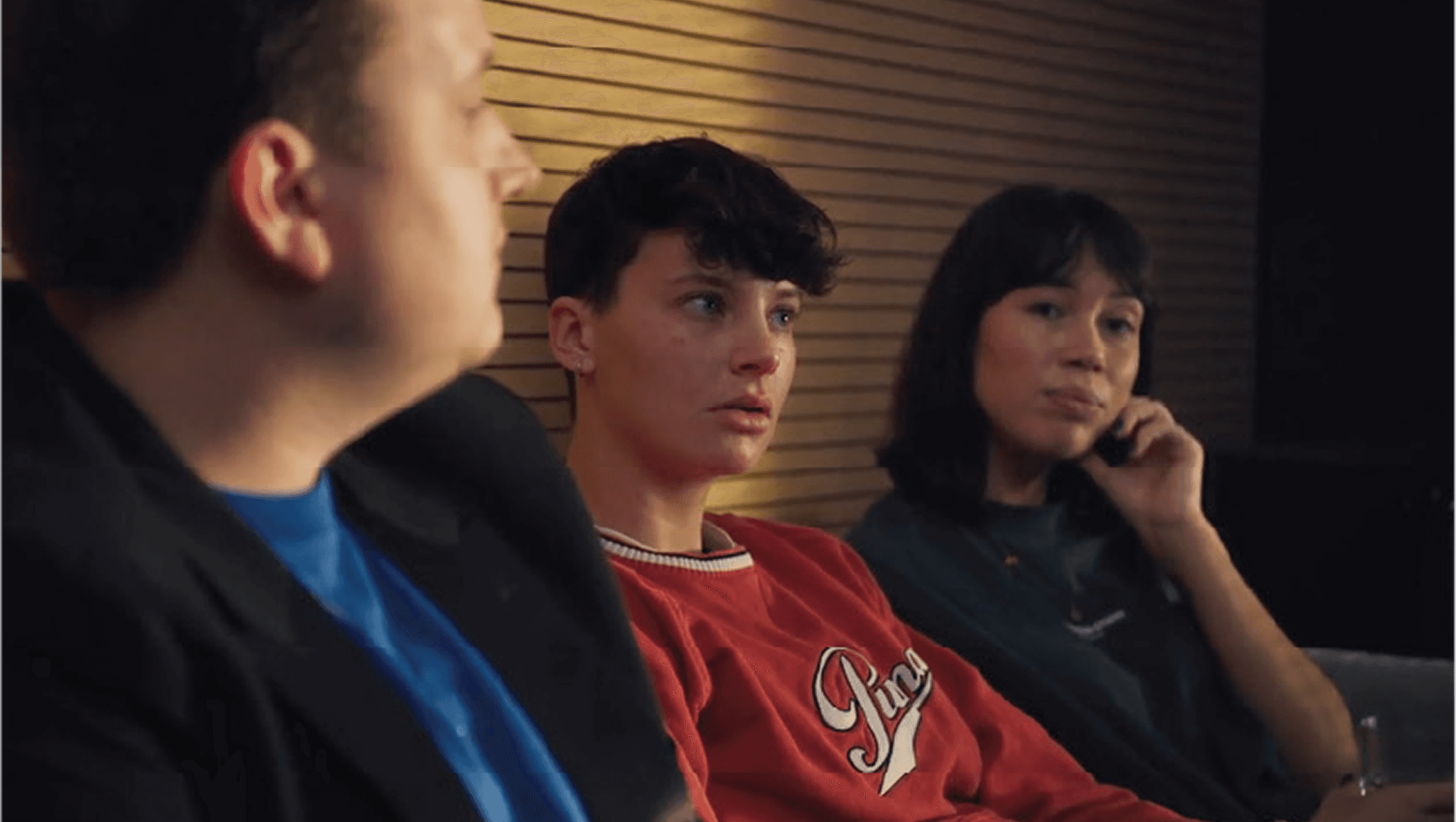 Three people seated before wooden slats: blurred man left, young person in red sweatshirt center looking ahead, woman right resting hand on face.