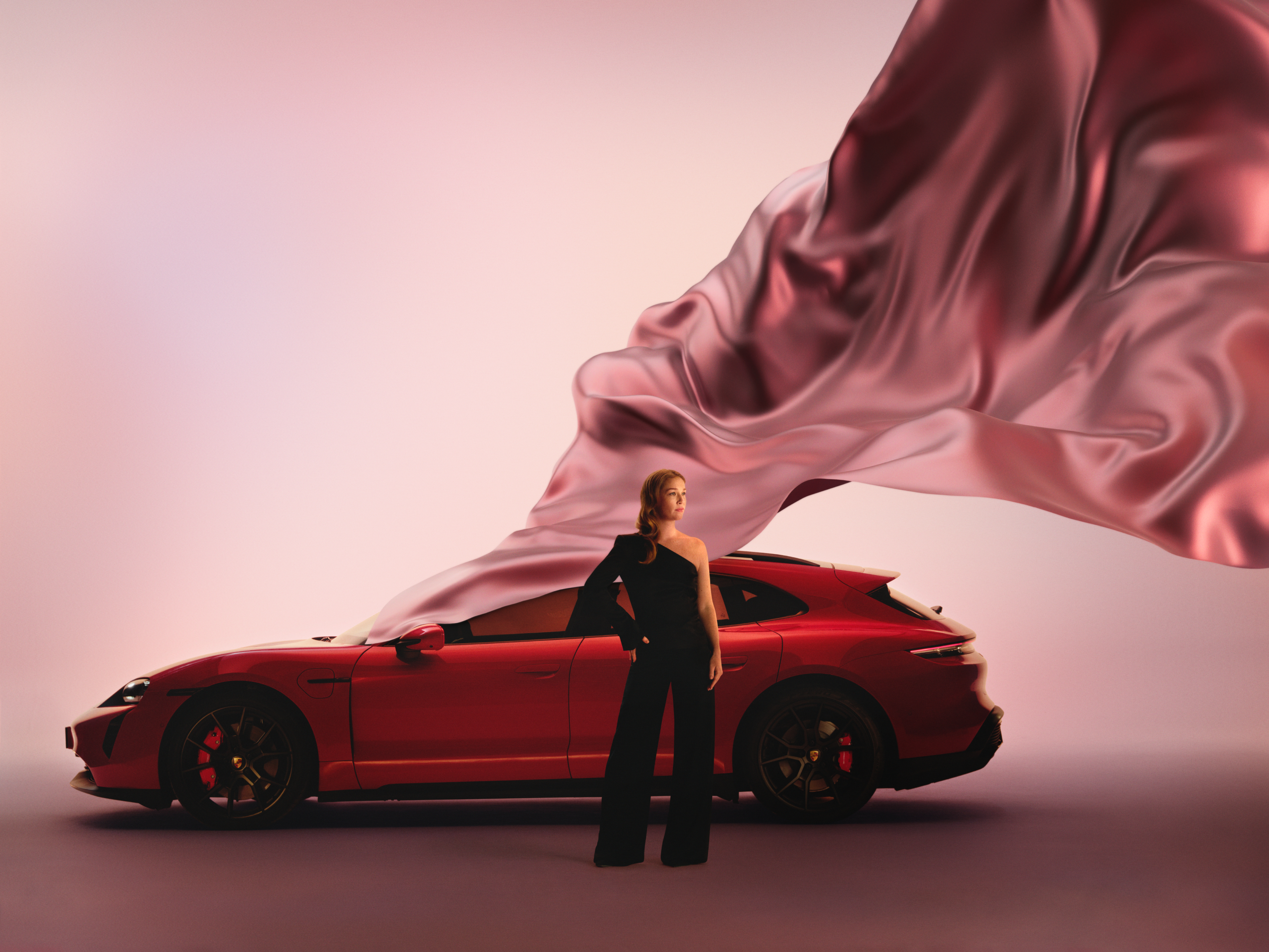 Woman in black outfit stands beside a red Porsche against a pink backdrop, large flowing satin fabric billowing overhead.