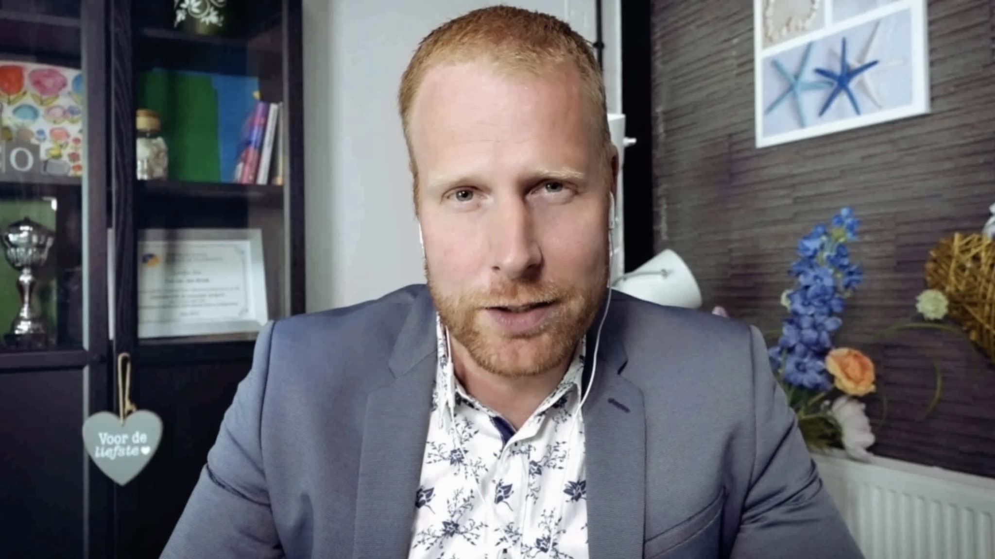 A medium shot of a light-skinned man with short reddish-blonde hair and a beard, wearing a gray blazer over a white floral patterned shirt. He is wearing wired earphones and looking directly at the camera against a home office background featuring a bookshelf, a framed certificate, and decorative floral wall art.