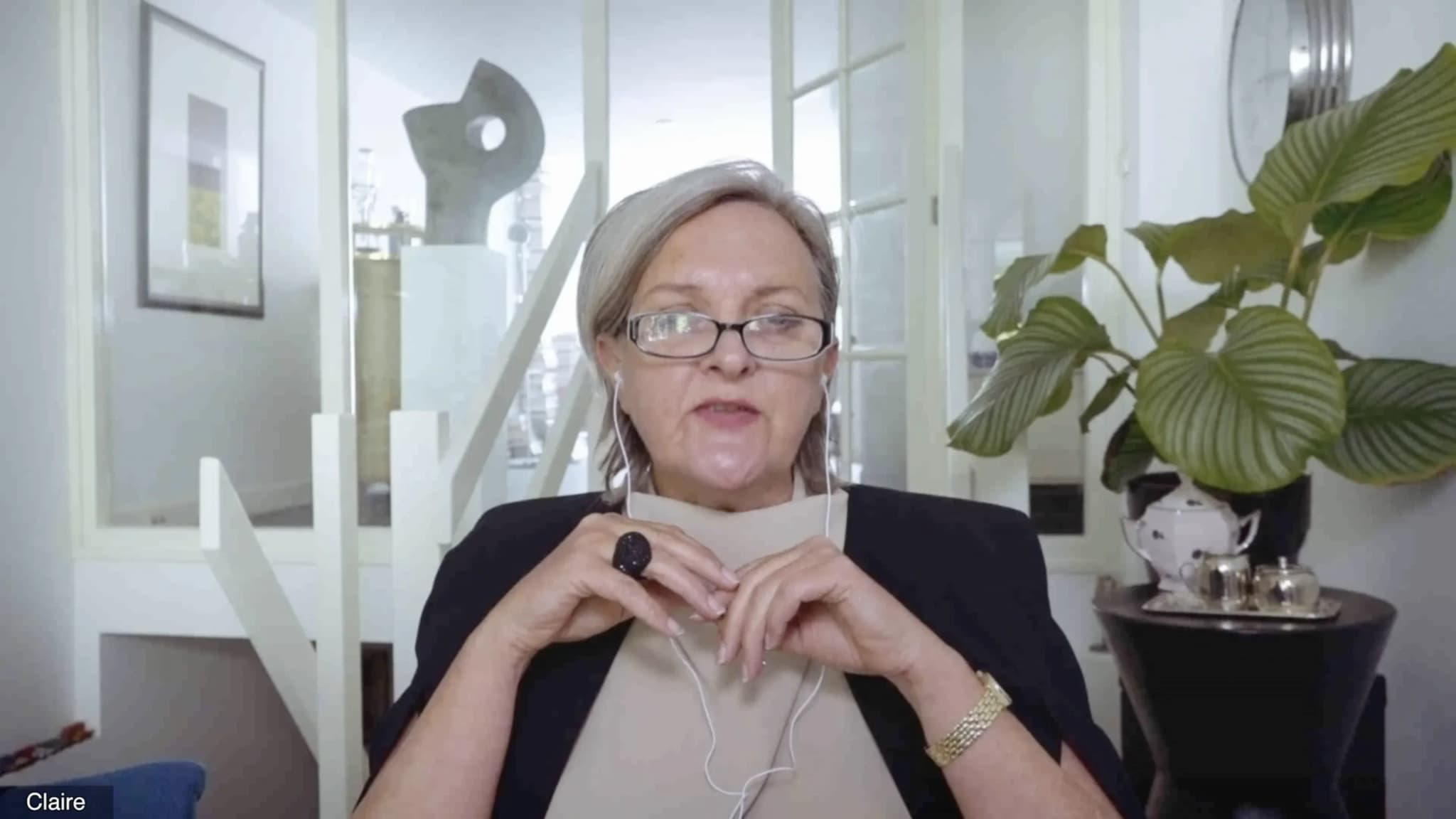 A medium shot of an older woman with short gray hair and glasses, wearing a black blazer over a beige top. She is positioned in front of a white staircase and a large green plant, with a silver tea set on a small table to her right. The name "Claire" is visible in a small tag at the bottom left, suggesting a video call setting.