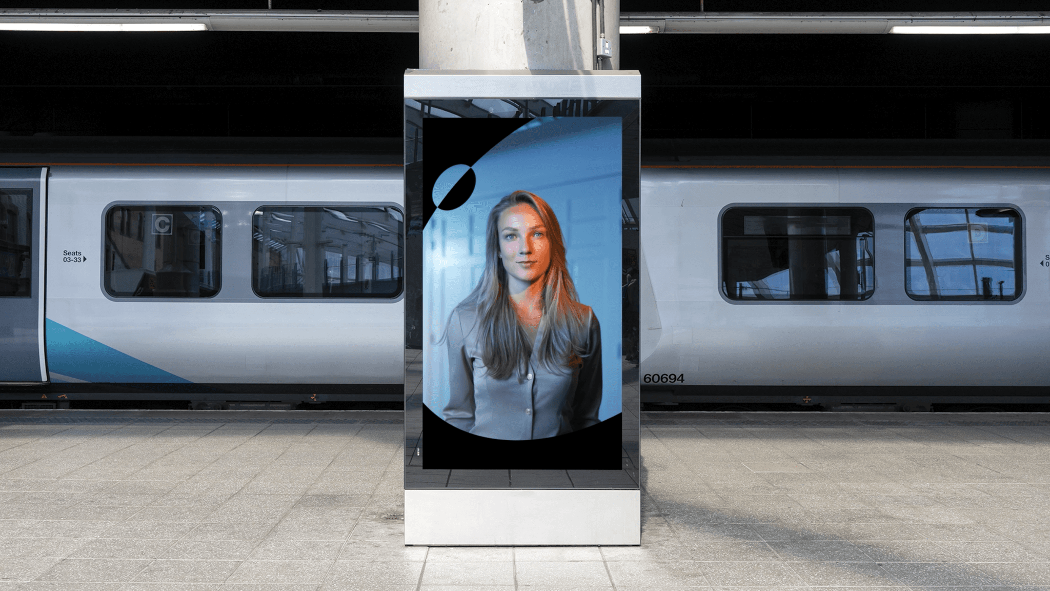 Vertical digital screen on a train platform showing a woman's portrait; train cars visible behind.