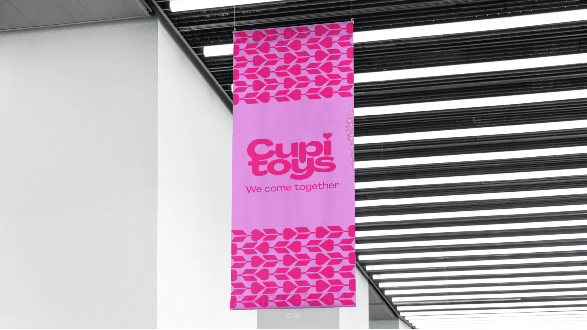 A vertical pink banner hanging from a ceiling in an indoor public space. The banner features the bright magenta "Cupid Toys" logo with the slogan "We come together" centered below it. The top and bottom borders of the banner are decorated with a repeating pattern of dark magenta stylized hearts and arrows.