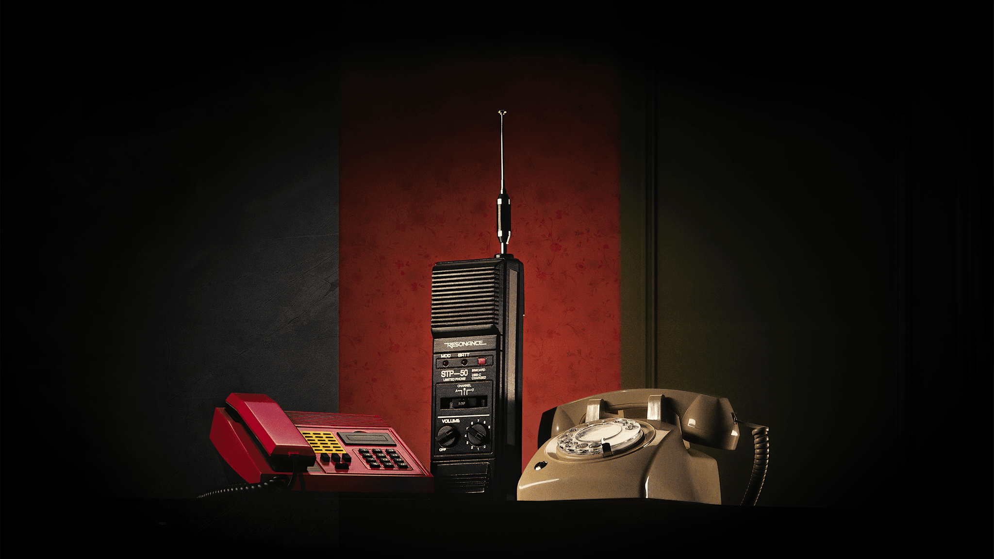 Three vintage phones on a table from different eras and Netflix shows: red push-button phone, tall black mobile with antenna, and beige rotary telephone against a dark backdrop.