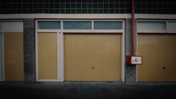 An exterior shot of a row of utilitarian garage doors made of yellow corrugated metal set into a gray brick building. A red drainage pipe and a small white utility box with red trim are visible on the brick wall between the closed doors.