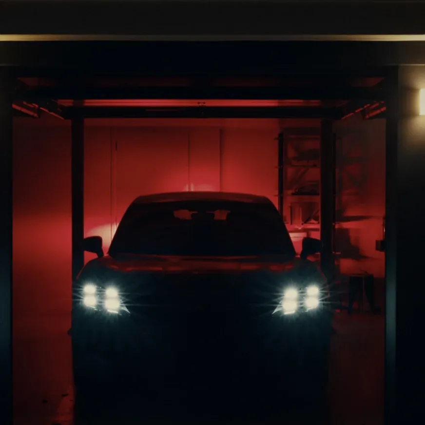 The front view of a dark Porsche inside a garage, illuminated by a dramatic red glow from behind. The car’s distinctive four-point LED headlights are brightly lit, casting sharp glares into the dark, industrial space.