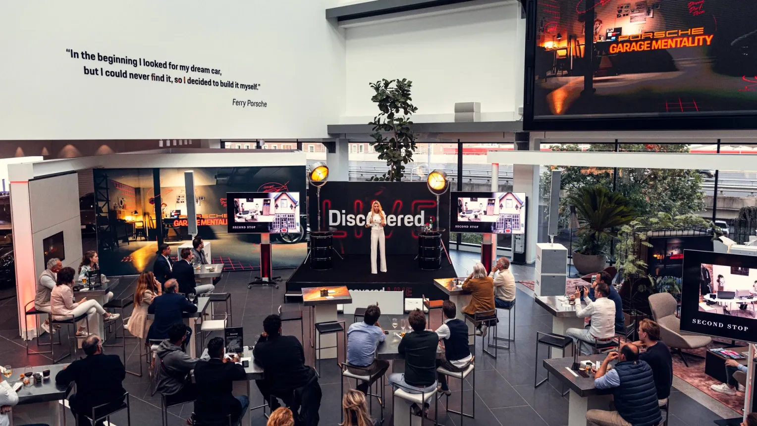 A wide shot of a Porsche event space featuring an audience seated at high tables facing a stage where a woman in white is speaking. A large wall quote by Ferry Porsche is visible on the left, and a massive screen displaying the "Porsche Garage Mentality" graphic hangs on the right.
