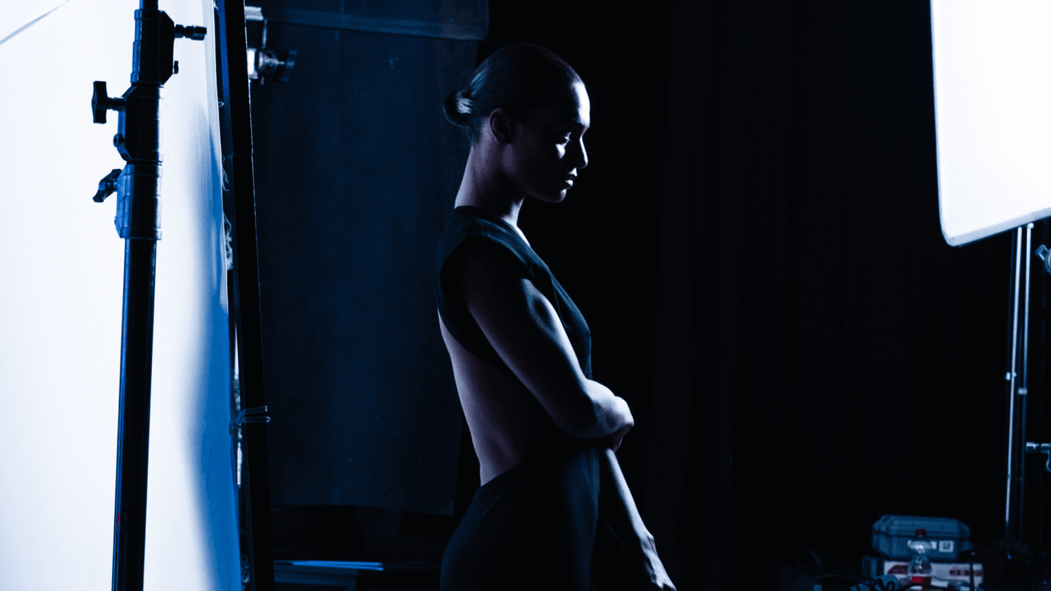 Profile of a woman in a backless dress standing in a dim photo studio, silhouette lit by softbox panels, arms crossed.