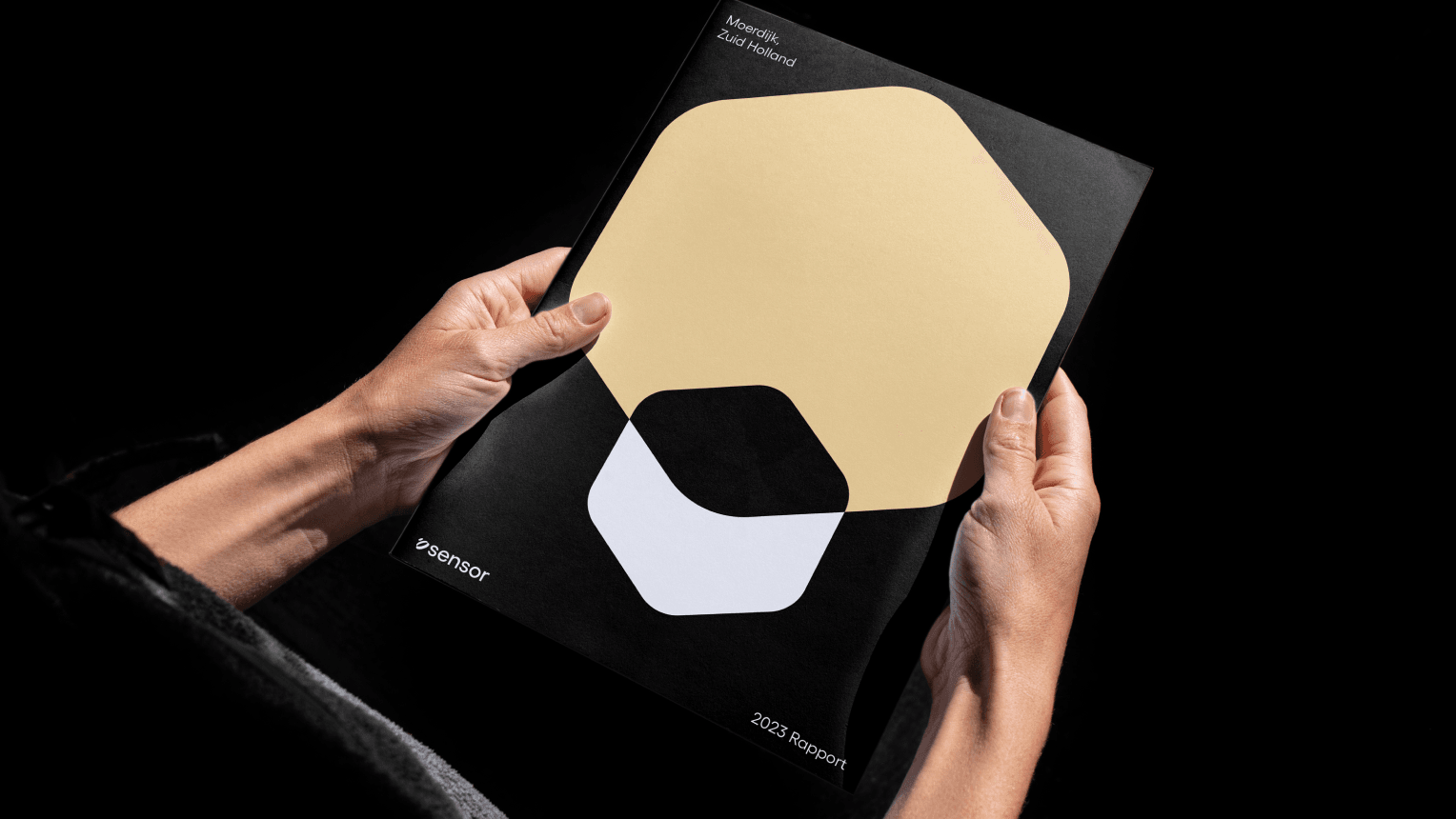 Two hands holding a black annual report with beige, black and white geometric shapes on the cover, photographed on a black background.