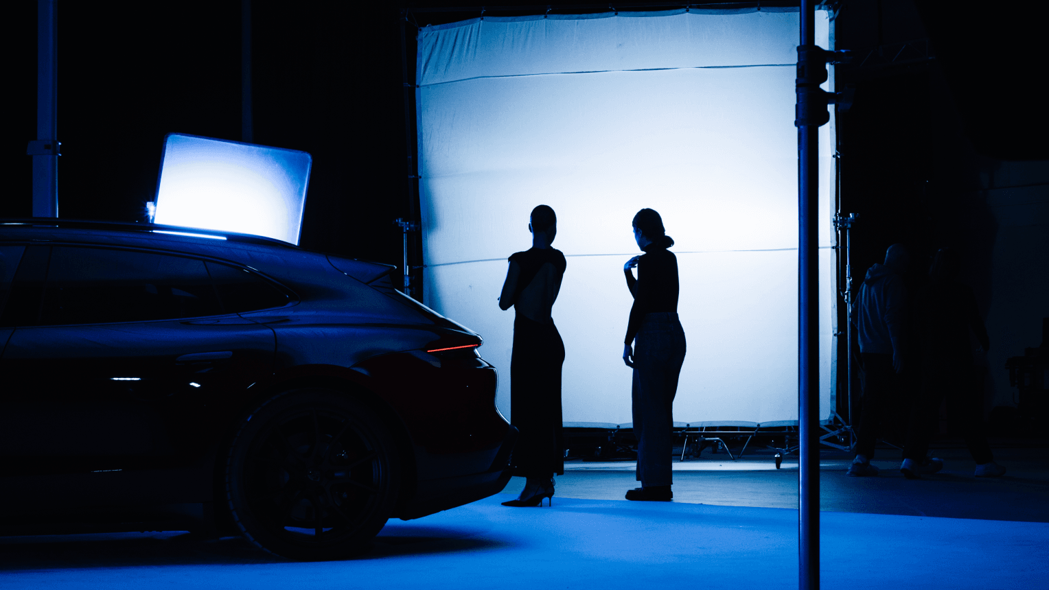 Silhouetted two people standing by an illuminated white backdrop beside a dark Porsche in a blue-lit studio.