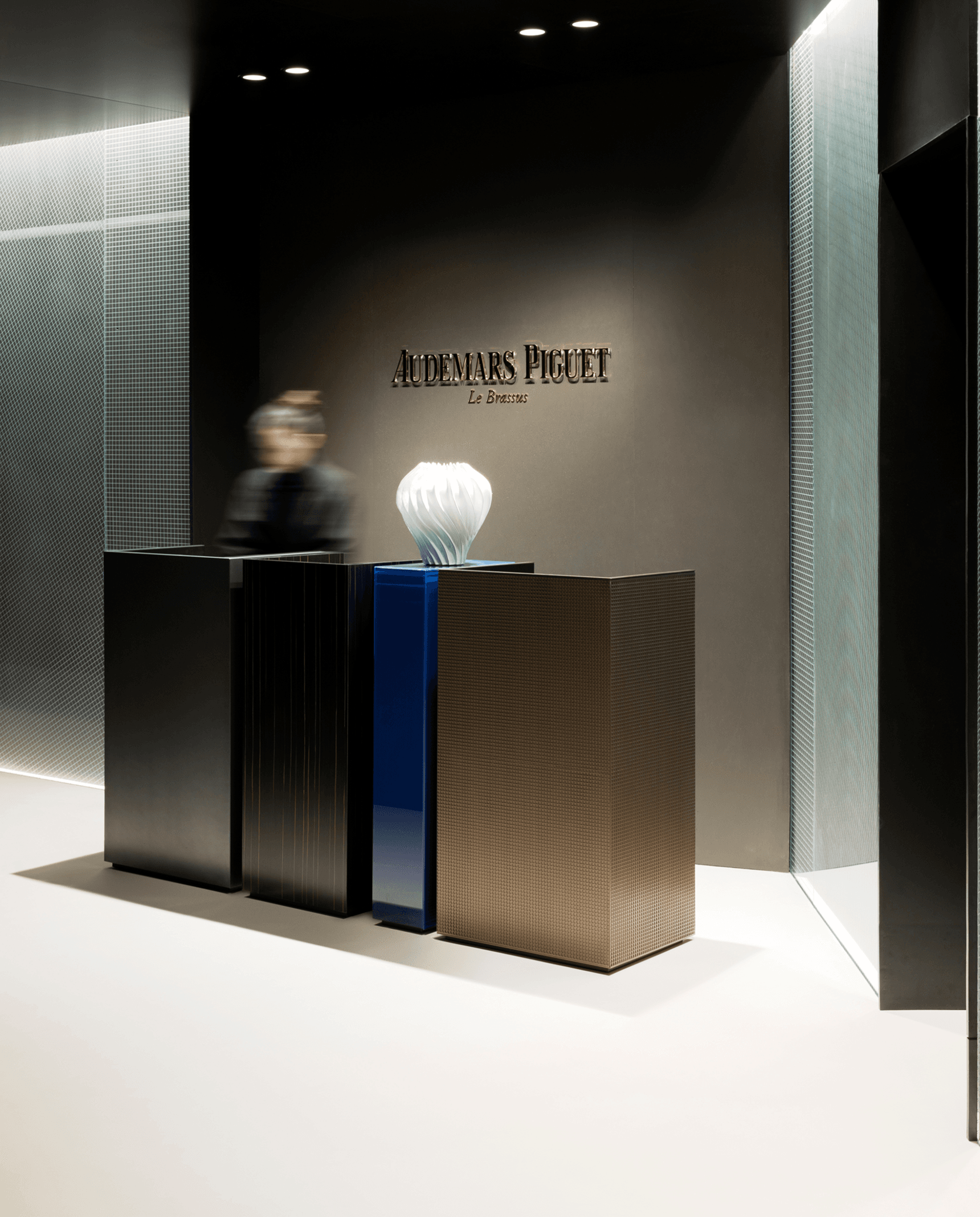 Minimalist Audemars Piguet reception: three dark podiums with a white sculptural lamp, blurred attendant and brand logo on a gray wall.