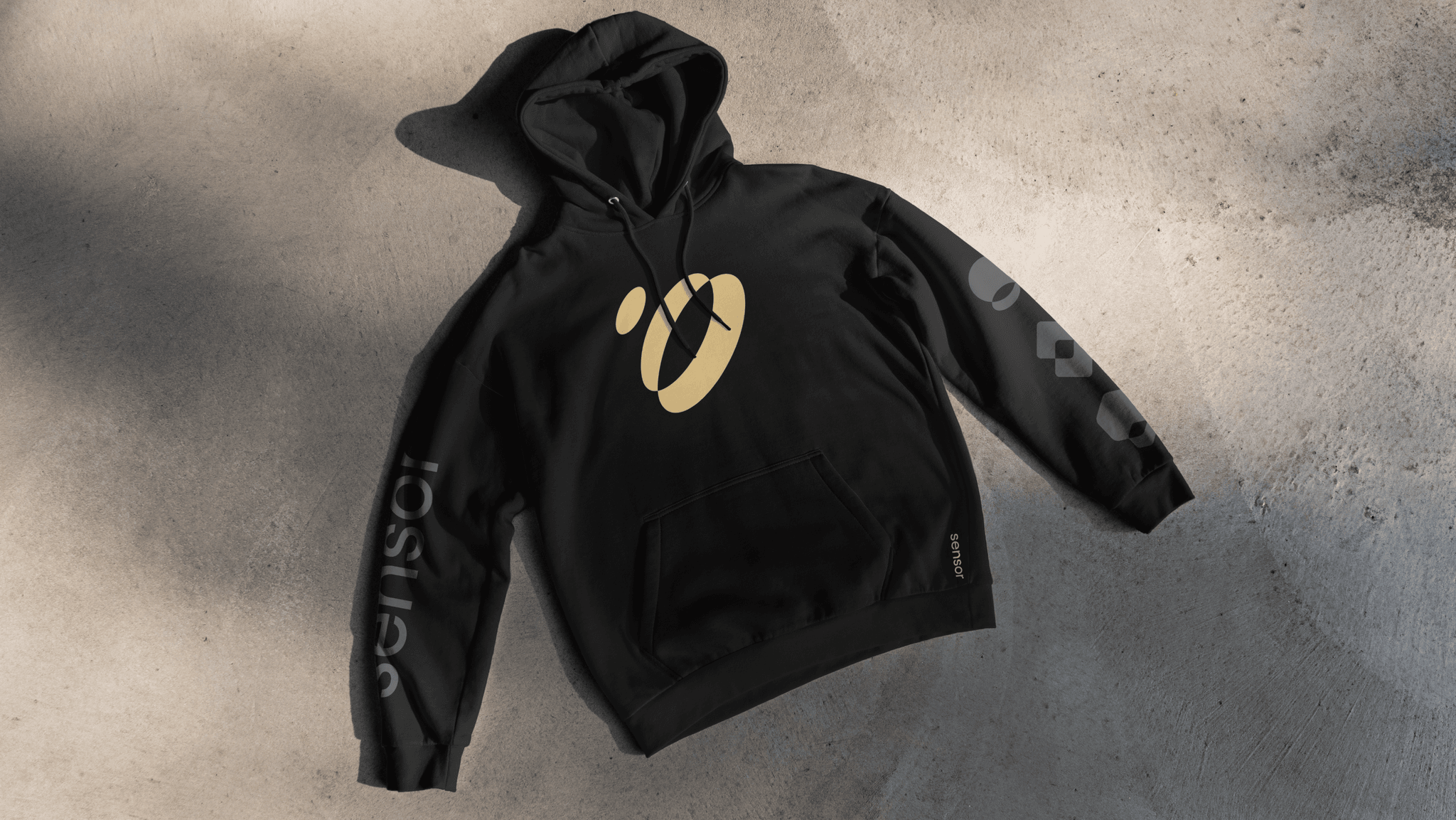 Black hoodie laid flat on concrete, yellow oval logo on chest and gray text/graphic running along the sleeves.