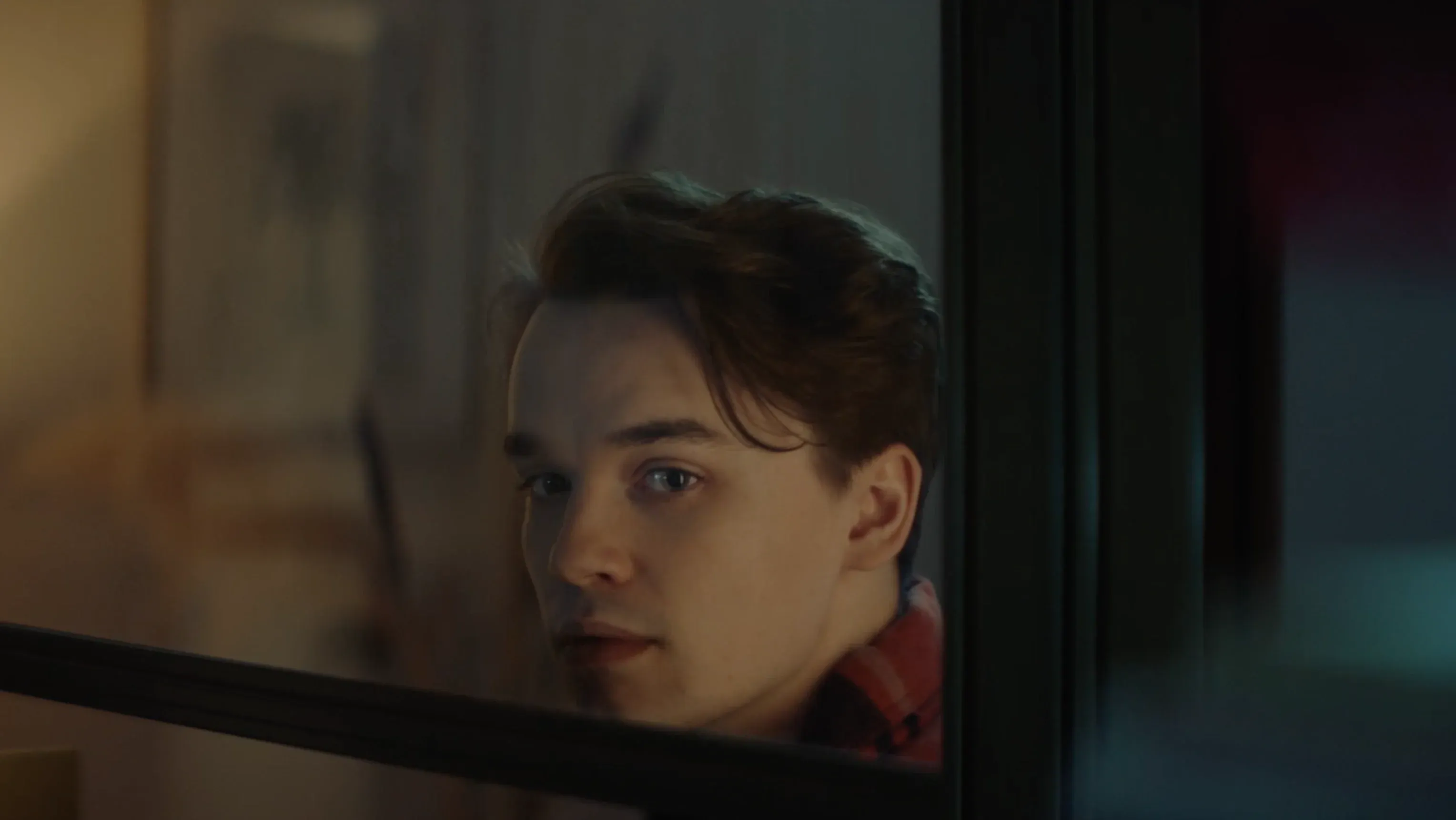 The same individual, now a young man, is seen in a more dimly lit, modern interior. He is wearing a very similar red and black flannel shirt, suggesting a bridge between his past and present self.