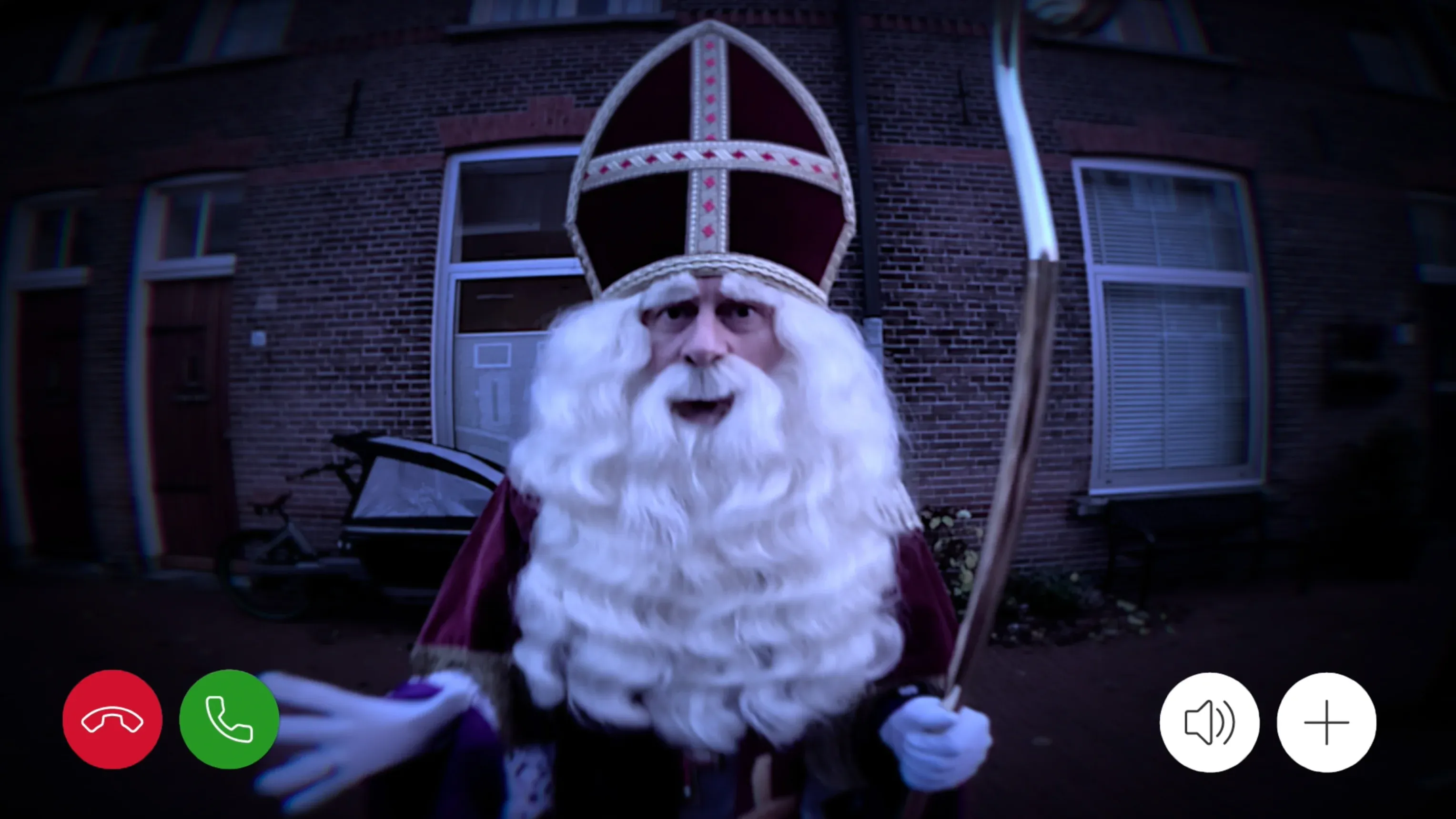 Doorbell-camera view: person in Sinterklaas costume with long white beard and staff, standing before brick row houses; call icons visible.