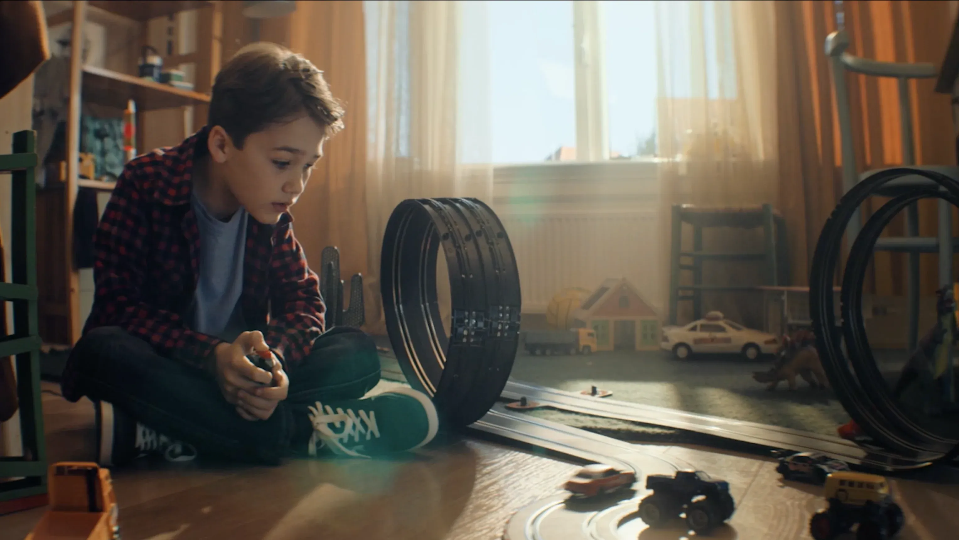 A young boy sits on a sunlit floor, intensely focused on a slot car racing set with loops and various toy vehicles. He wears a red and black checkered flannel shirt over a light blue t-shirt and dark jeans.