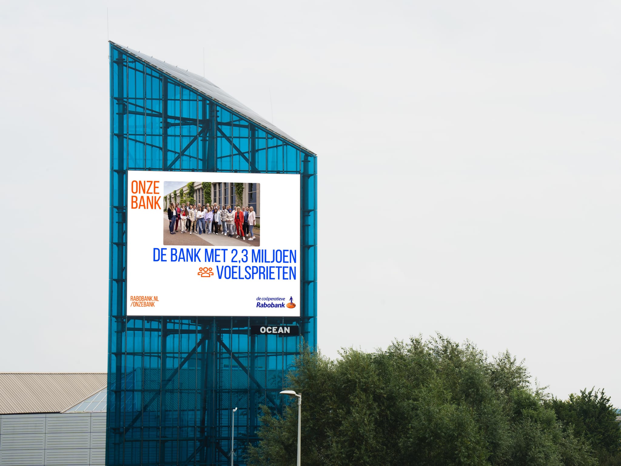 Blue glass tower with large digital billboard showing a Rabobank ad; trees in foreground under cloudy sky.