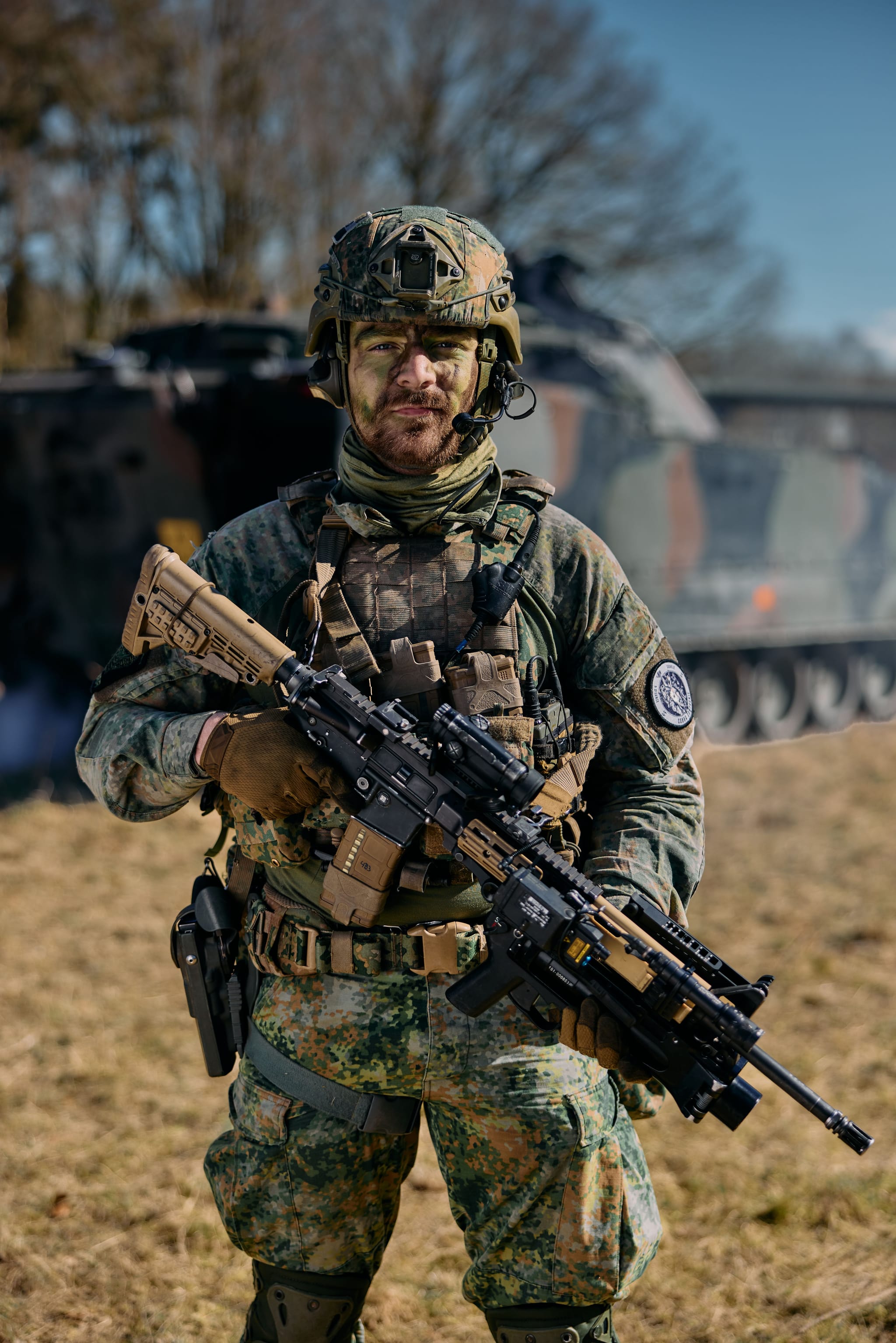 Camouflaged soldier with helmet and face paint holding a rifle, standing in a field in front of an armored vehicle.