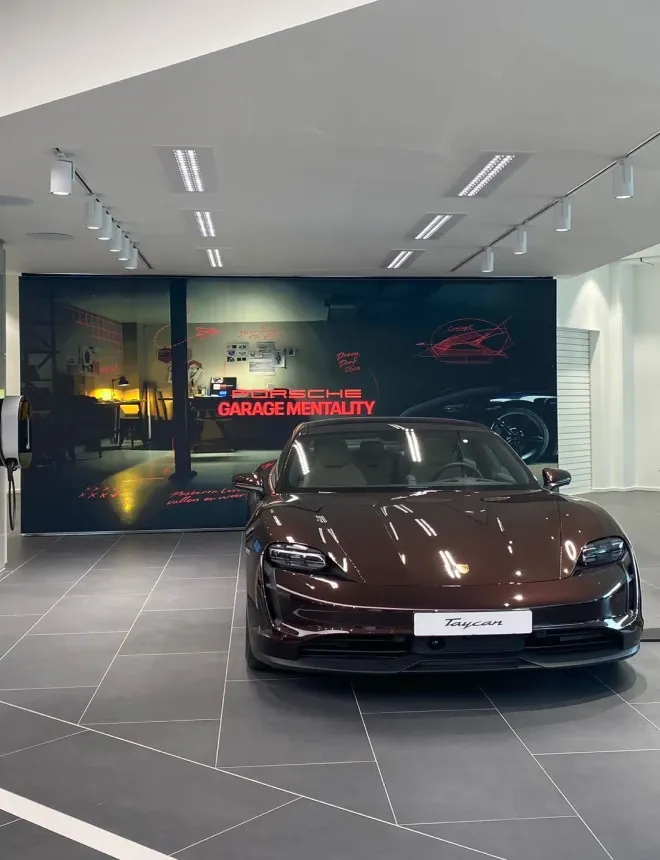 A bronze Porsche Taycan is parked in a modern, brightly lit showroom with a tiled gray floor. In the background, a large floor-to-ceiling banner displays the dark, neon-sketched garage from the "Porsche Garage Mentality" campaign.