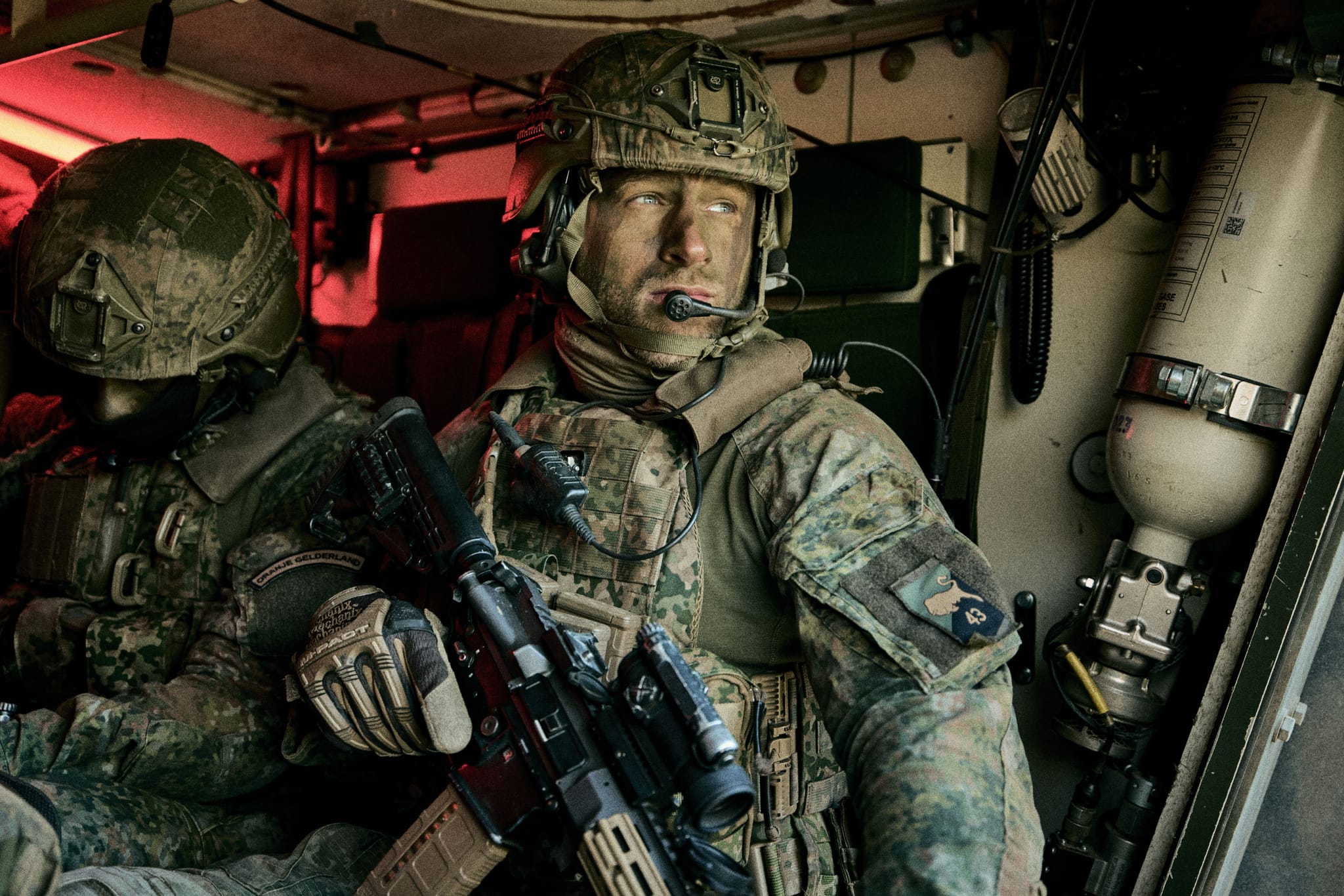 Soldiers in armored vehicle; foreground soldier wearing helmet and headset, holding a rifle and looking out.