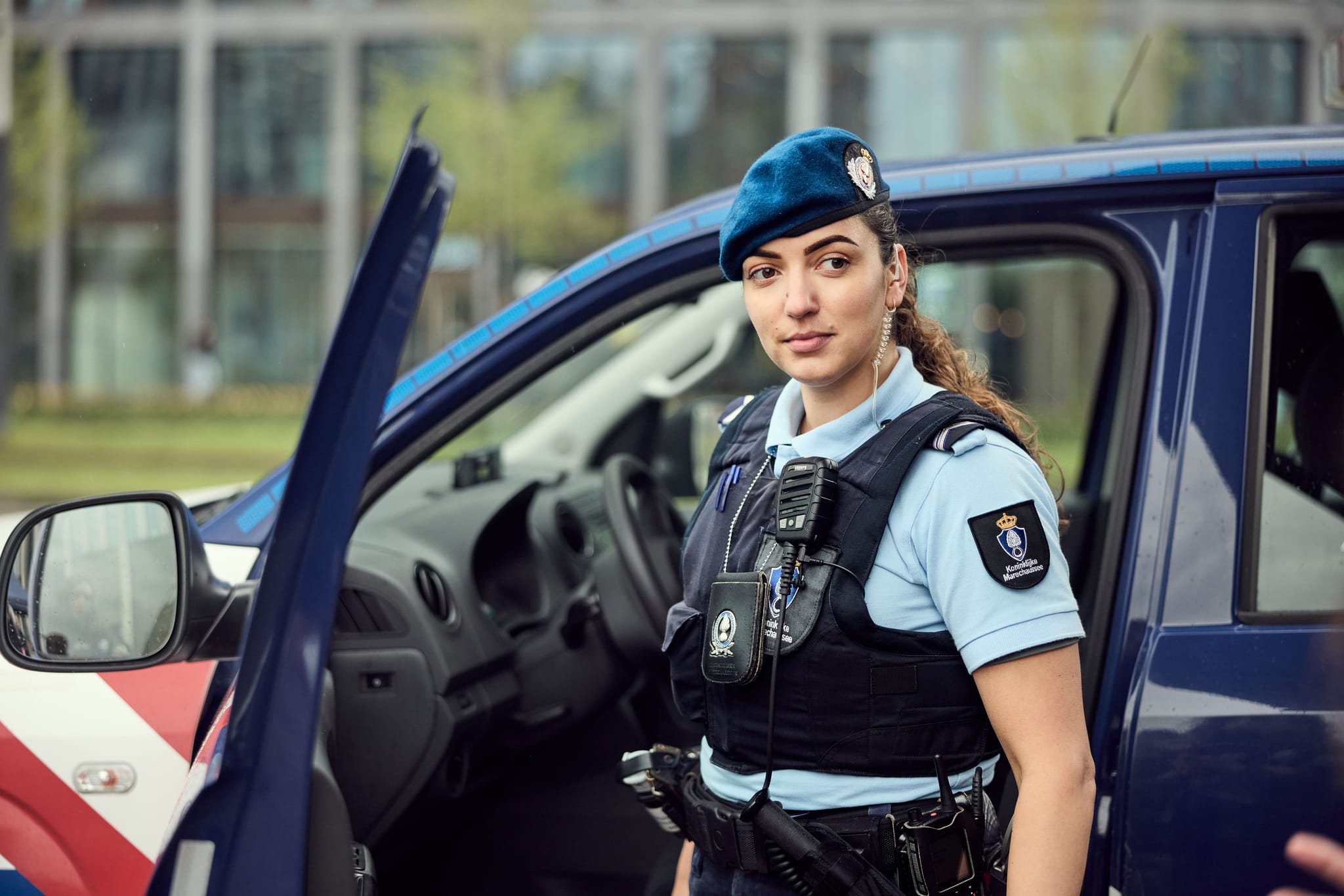 Female police officer in blue beret stands by an open patrol car door, wearing a tactical vest and radio.