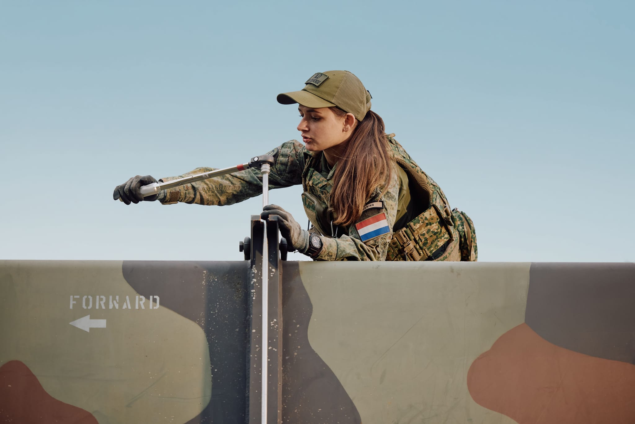 Soldier in camouflage uniform and cap, wearing gloves, tightening a metal latch on a painted military barrier marked "FORWARD."