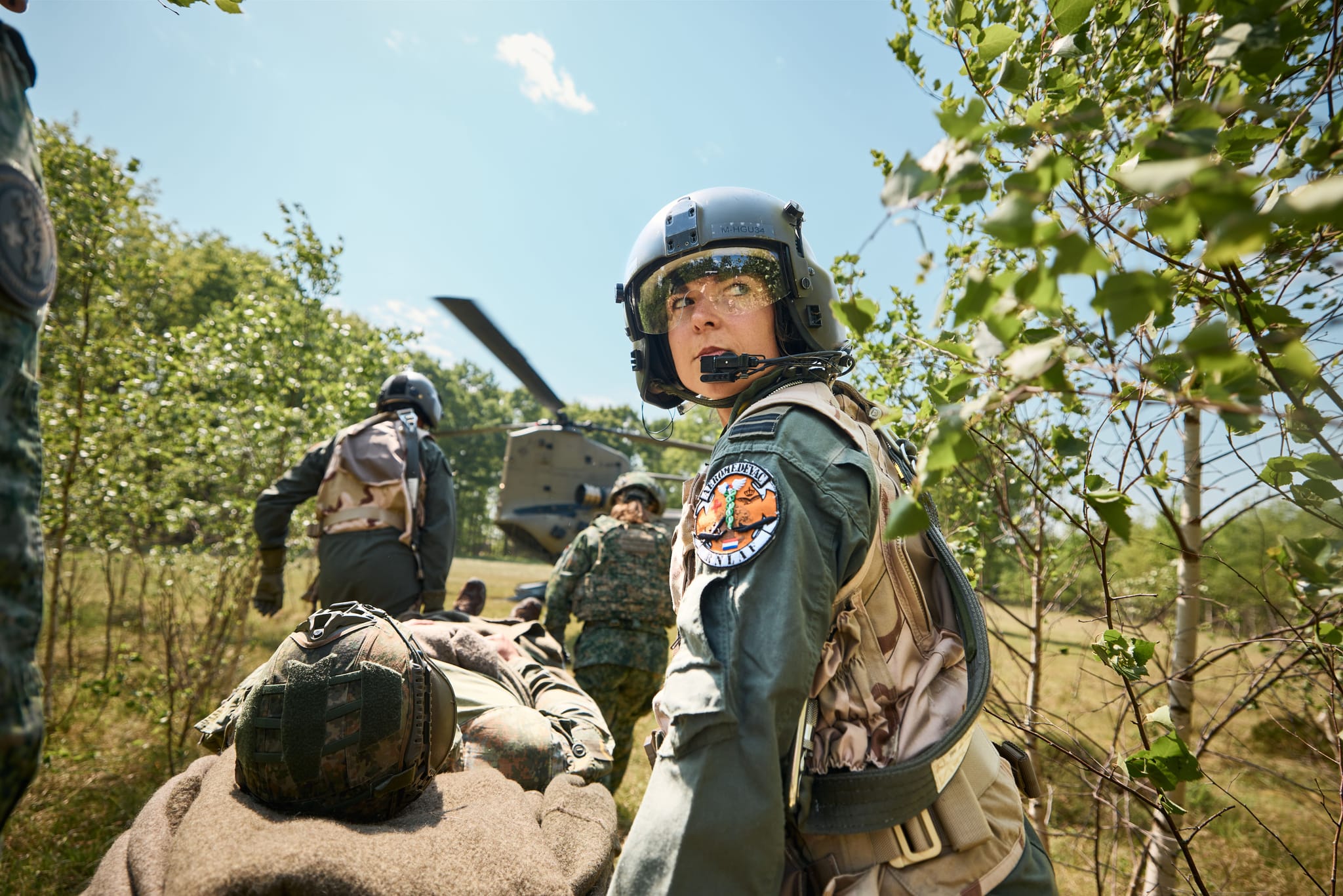 Helmeted military medic looks back as teammates carry a stretcher toward a helicopter in a wooded clearing.