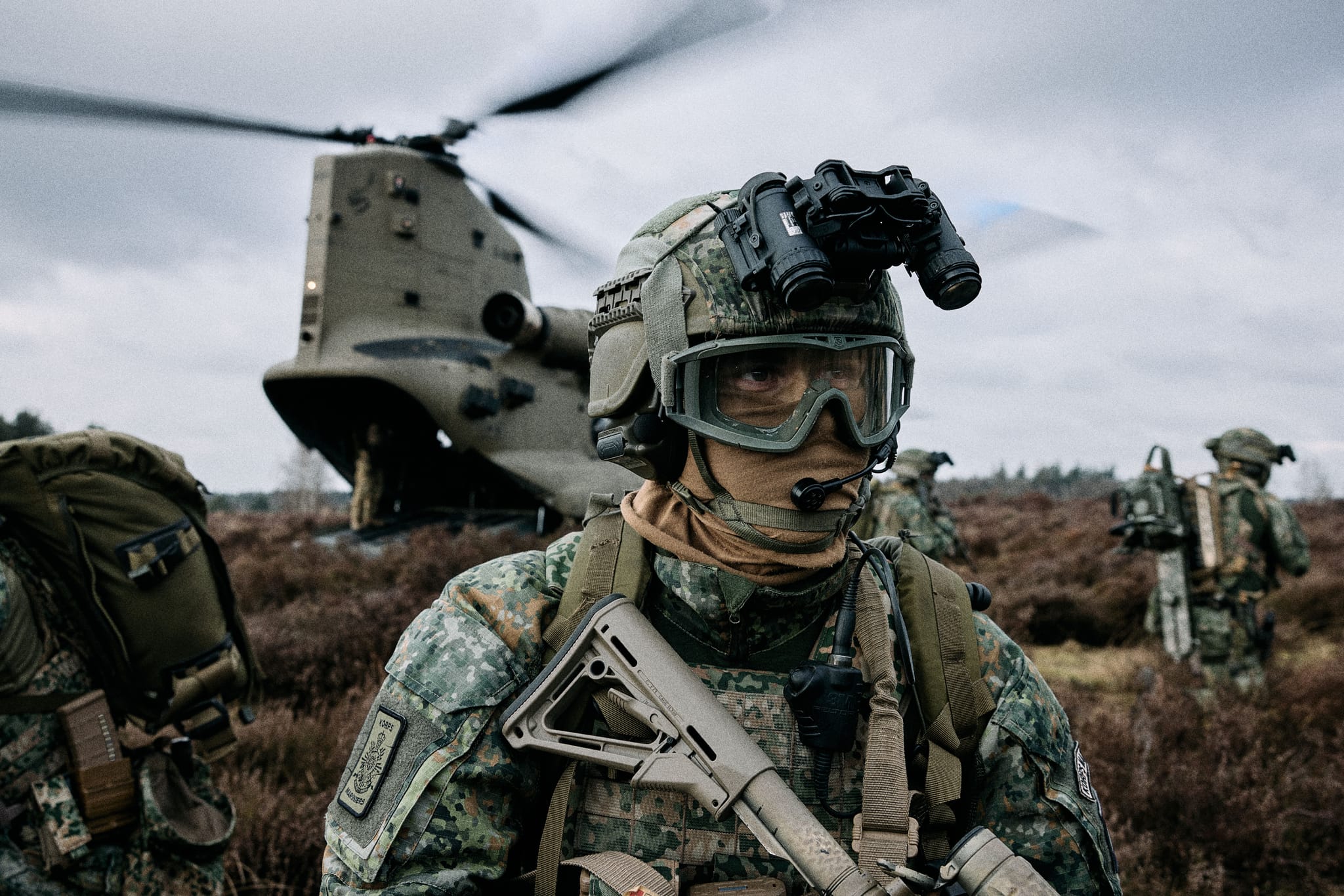 Soldier in digital camouflage with helmet-mounted night-vision and rifle, Chinook helicopter landing behind on open moorland.