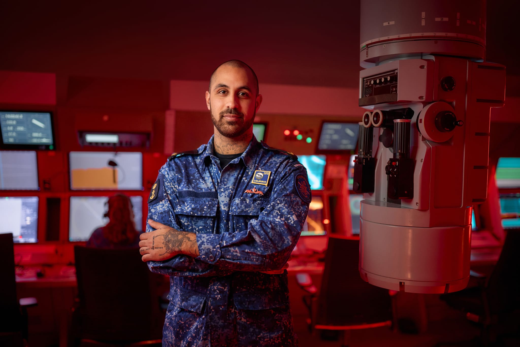 Man in blue digital camouflage uniform stands with arms crossed in a red-lit control room beside a large imaging machine and monitors.