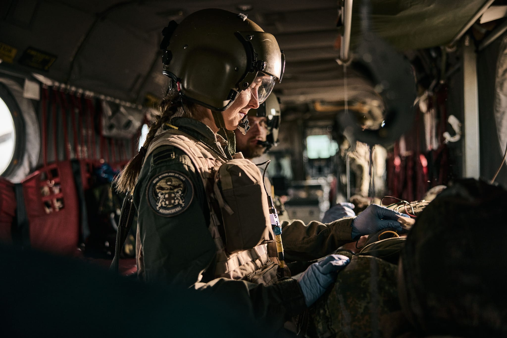 Military medic wearing a helmet and gloves tending to an injured soldier on a stretcher.