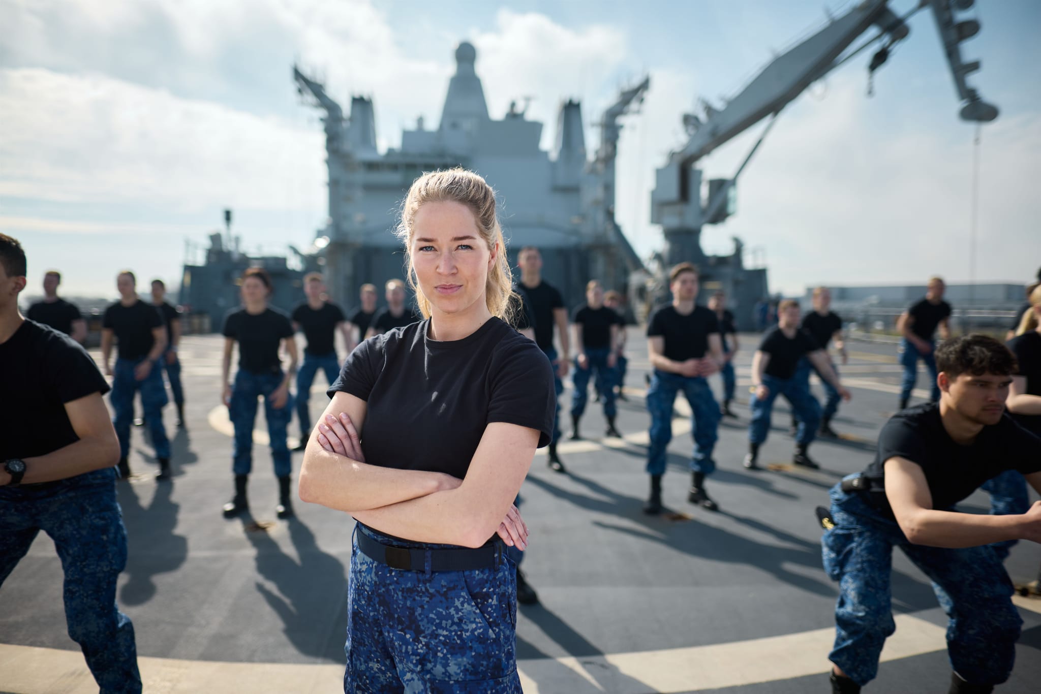 Female sailor in black shirt and blue camouflage pants stands with arms crossed on a ship deck, crew exercising behind her.