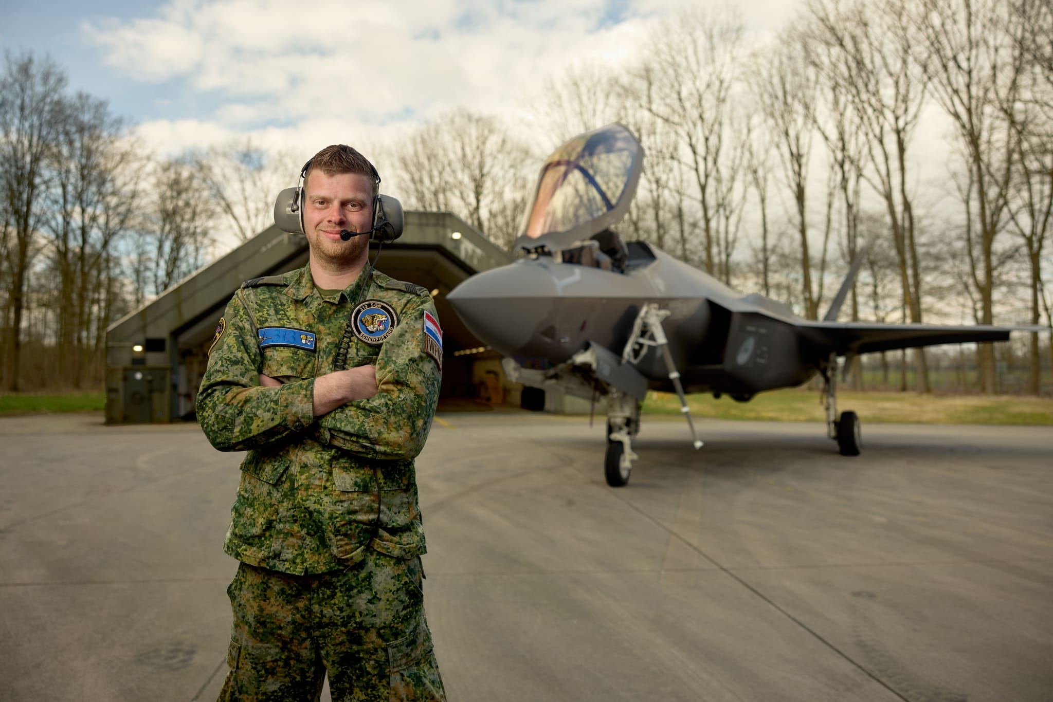 Camouflaged uniformed pilot with headset, arms crossed on runway in front of a stealth fighter jet and open hangar.