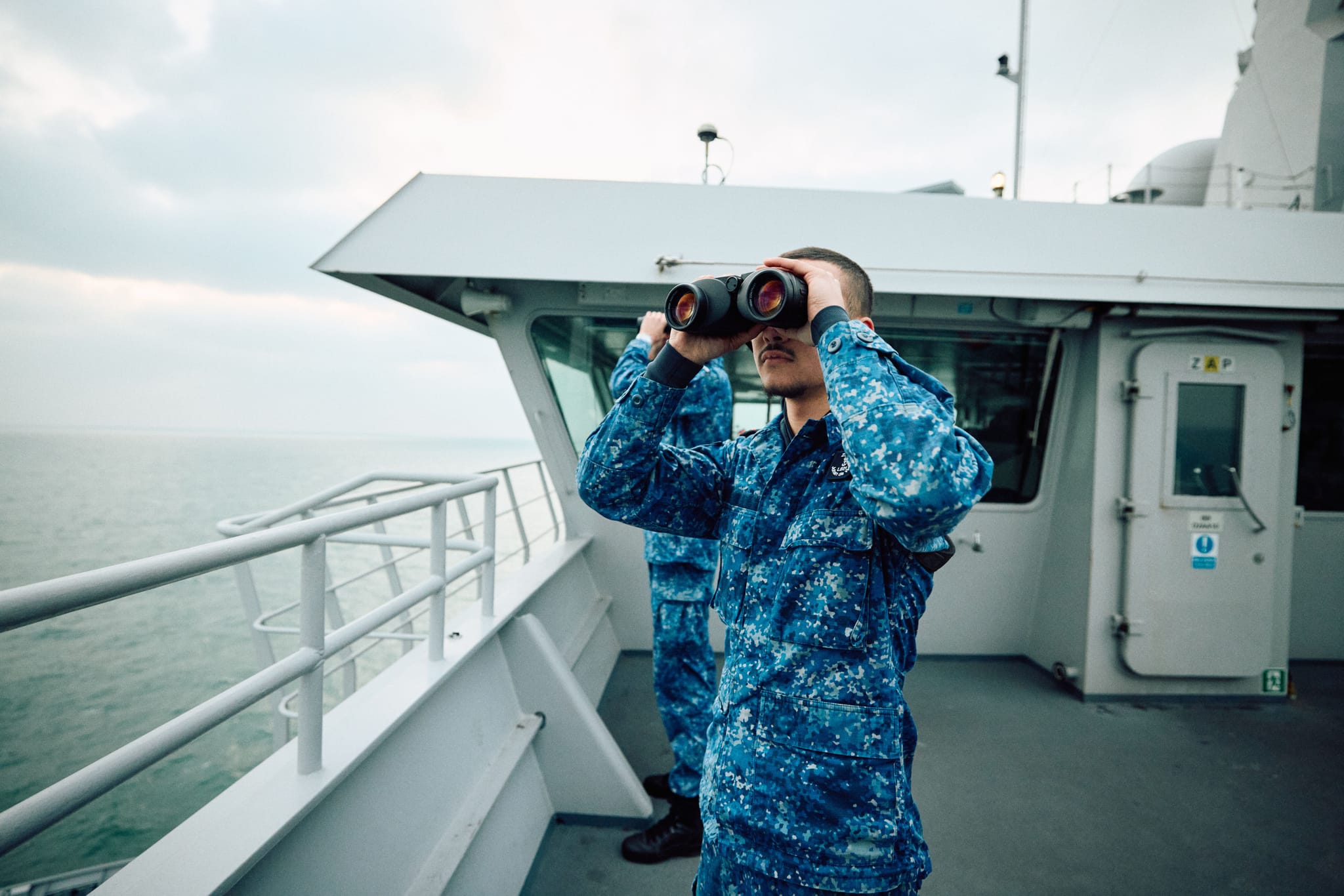 Sailor in blue digital camouflage using binoculars on a ship deck, another crew member and the sea horizon in the background.