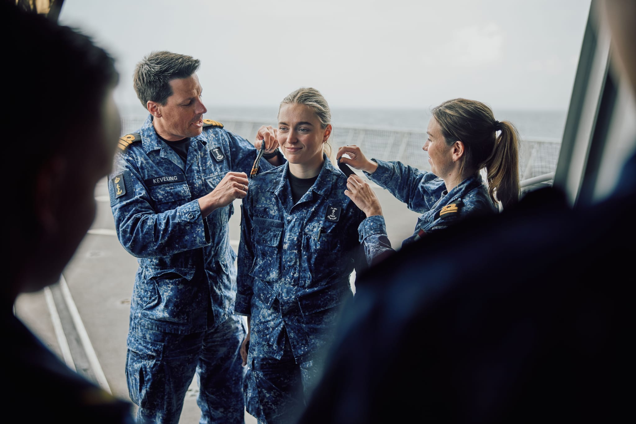 Two sailors pin rank insignia on a smiling female sailor aboard a ship, colleagues watching with the sea in the background.