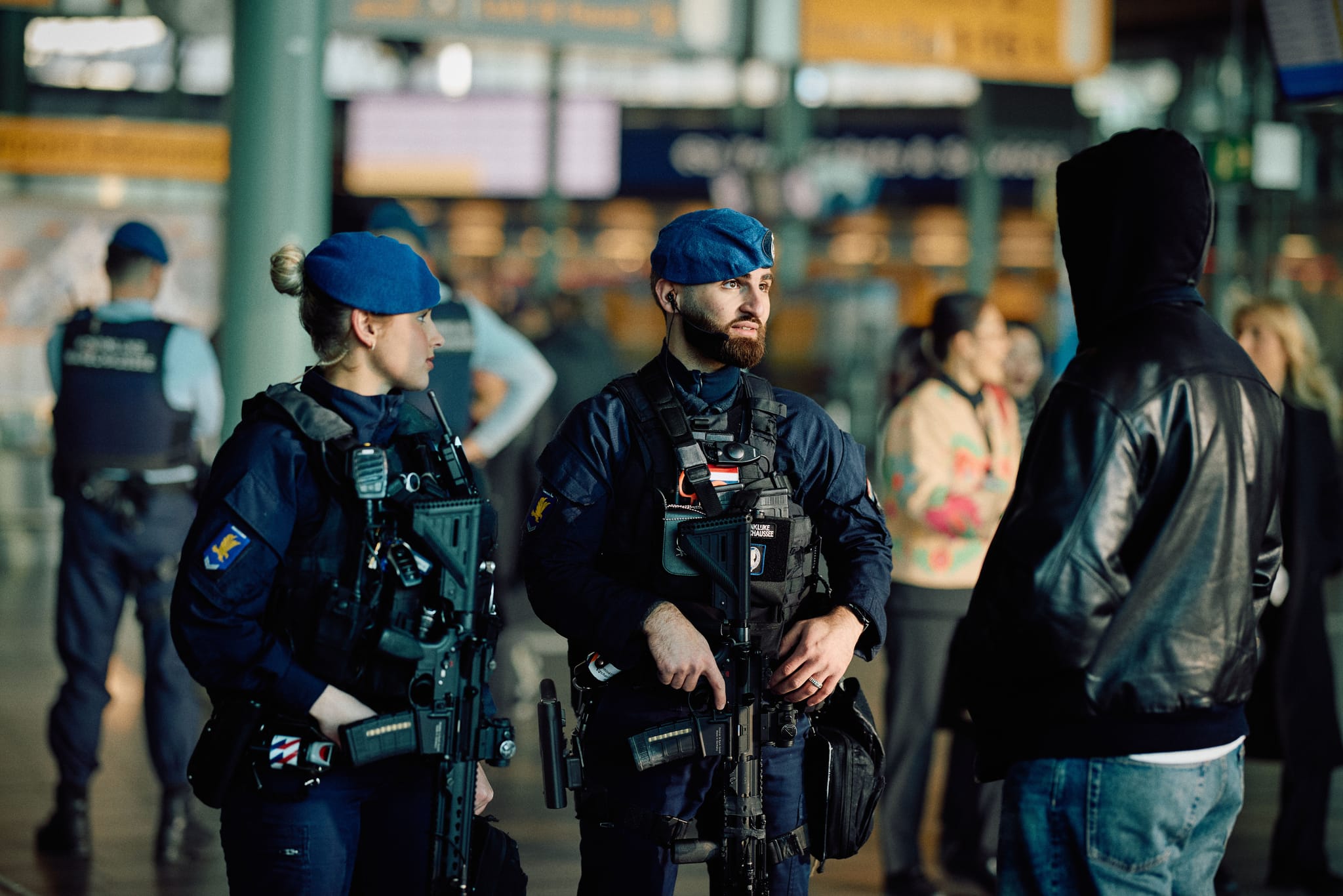 Two armed police officers in blue berets talk with a hooded man in a crowded train station concourse.