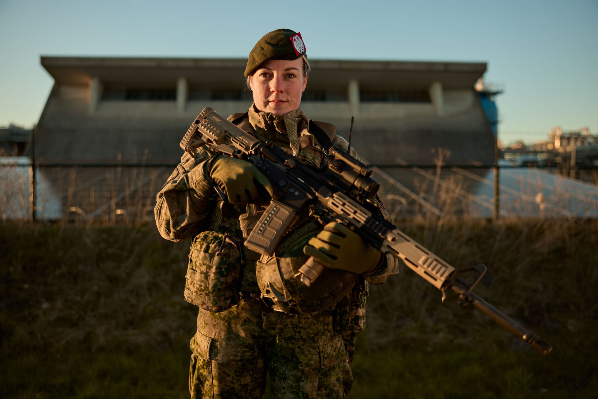 Armed soldier in camouflage and green beret holding a rifle, standing outdoors in front of a concrete industrial building.